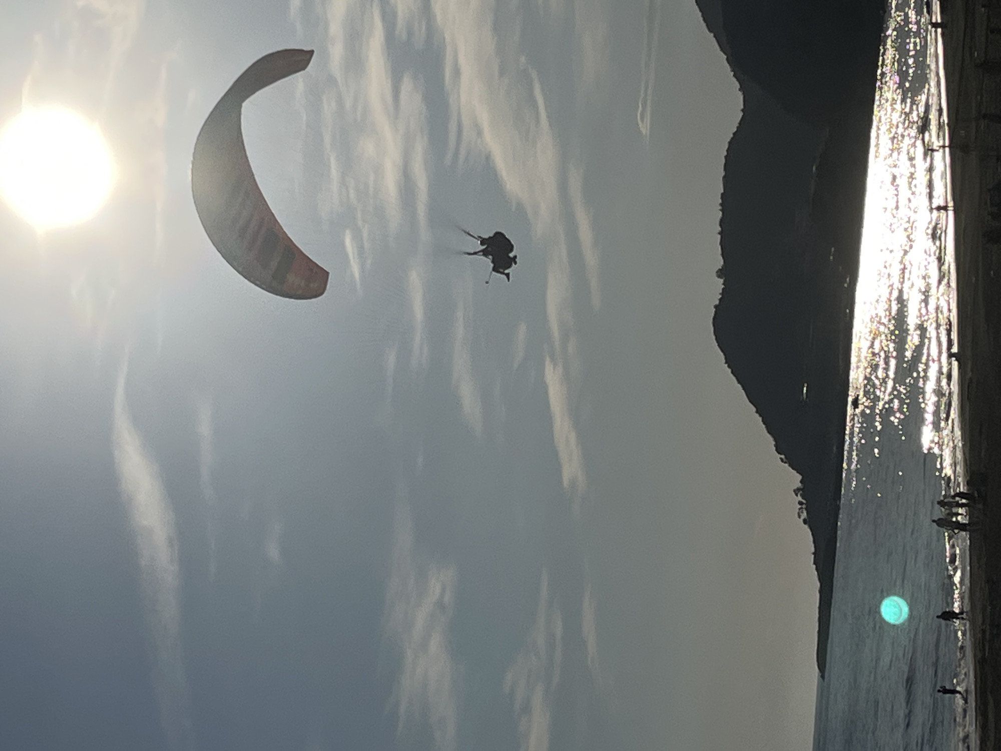 The image depicts a paraglider in flight above a beach, likely in Alanya, Turkey. The sun is positioned in the upper left corner, obscured by clouds, casting a bright light across the sky. A paraglider, with a red and white canopy, is visible in the air, along with two indistinct figures suspended below. The beach runs along the right side of the frame. There are other, blurry figures present on the beach. A dark, mountainous coastline extends to the edge of the beach. The water is reflecting the sunlight.
