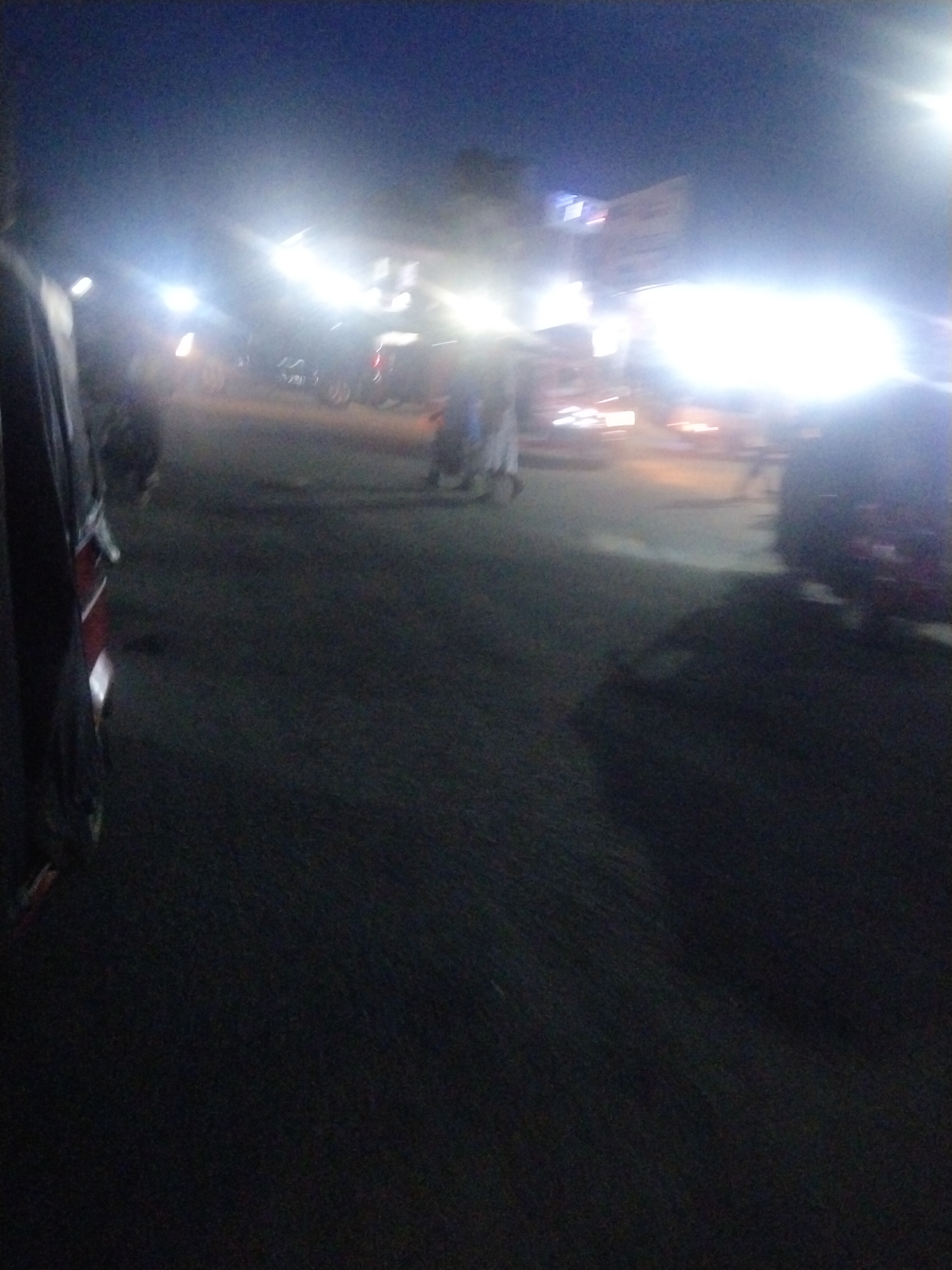 The image depicts a highly blurry and dark night scene on a street, likely in an urban or semi-urban environment. The primary subjects are numerous bright, overexposed artificial light sources, presumably from vehicle headlights, streetlights, and possibly illuminated buildings or signs. These intense lights create significant glare and lens flare, obscuring much of the detail.
In the immediate foreground on the left, a portion of a vehicle, displaying red and dark colors, is visible, suggesting the perspective is from within or very close to another vehicle. The ground beneath is a dark, indistinct surface, likely a road or paved area.
Faint, silhouetted forms of several people can be vaguely discerned in the middle ground, appearing to walk or stand amidst the vehicles. Their features and specific activities are entirely obscured by the blurriness and low light conditions. The presence of multiple light sources and indistinct forms suggests general traffic and movement on a busy street. No legible text is visible due to the image quality and overexposure, though some faint markings might exist on distant, illuminated structures. The overall impression is that of a bustling street scene in Uyo, Nigeria, after dark, characterized by a lack of clarity and an abundance of bright, diffused light.