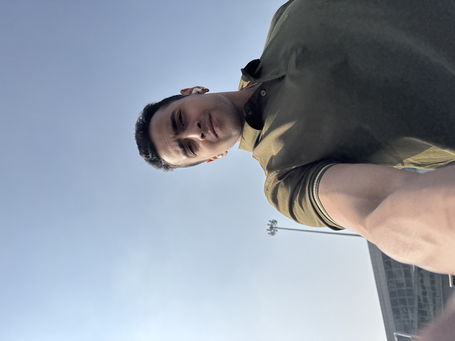 A man in an olive green shirt is seen from a low angle, tilting the camera toward himself. Clear blue sky fills the background, with a light pole and a tall building visible in the distance. His face is partially shaded, revealing dark hair and a neutral expression. A white earbud is visible in his ear. His arm is extended toward the camera, showing a hint of muscle definition.