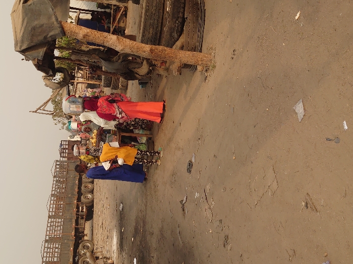 The image depicts an unpaved outdoor area in Ajaokuta, Nigeria, characterized by dry, light-brown earth and scattered debris including paper and plastic. In the midground, a group of approximately six individuals is gathered around a low wooden vendor stall displaying yellow and green produce. One person behind the stall, wearing a white head covering, attends to the goods. Among those facing the stall are an individual in a long blue garment, a young person in a yellow top and patterned leggings, and another individual in a reddish-orange patterned dress with a matching head covering, all seen from behind. Two individuals further back are observed carrying containers balanced on their heads. In the background to the left, two dark-colored, multi-axle flatbed trucks with metal railing structures are parked. To the right, a makeshift shelter constructed from a tree trunk and a dark fabric tarp stands near a stack of used vehicle tires.