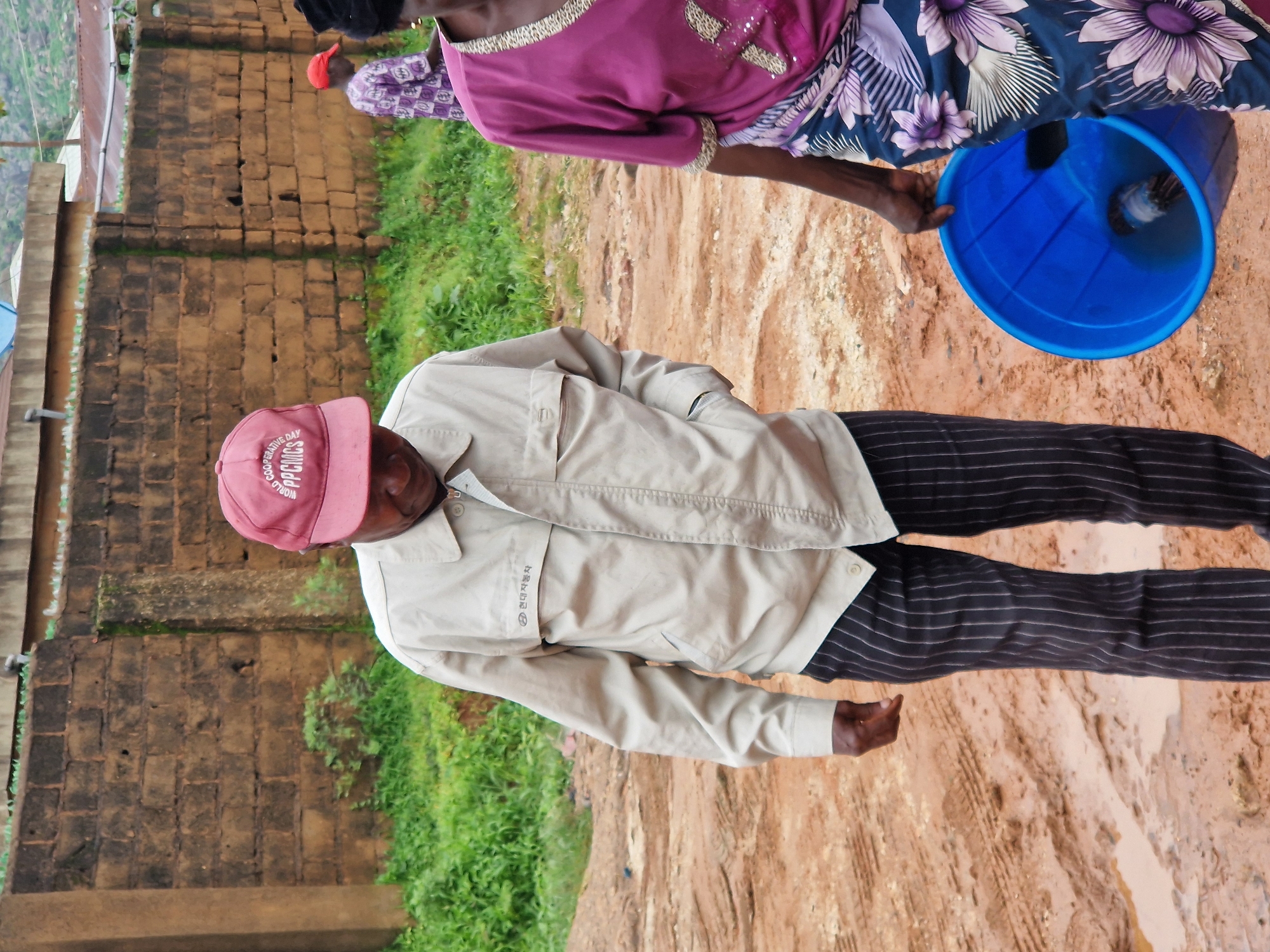 In Kabong, Nigeria, a man wearing a light-colored jacket and pinstripe pants stands in a muddy, uneven outdoor area. He has a red baseball cap on his head with the words "WORLD COOPERATIVE DAY PPCNMCS" embroidered on it. To his right, a woman in a purple top and a patterned skirt is visible, carrying a bright blue bucket. In the background, a brick wall and lush green vegetation are present.
