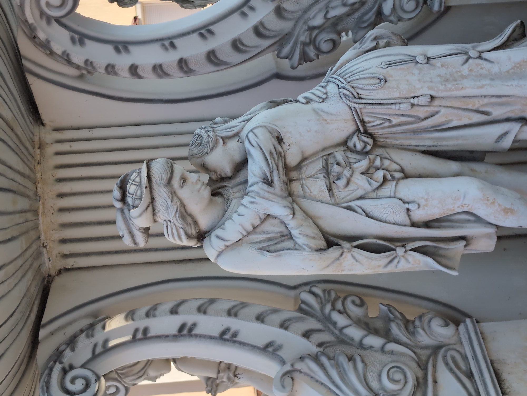A low-angle, close-up shot of a white, likely marble, sculpture depicting a man and woman, presumably lovers, in what appears to be a tender moment. The man wears a cap. The sculpture is adorned with ornate carvings. It is located at the Alexander Nevsky Cathedral in Izhevsk, Russia.