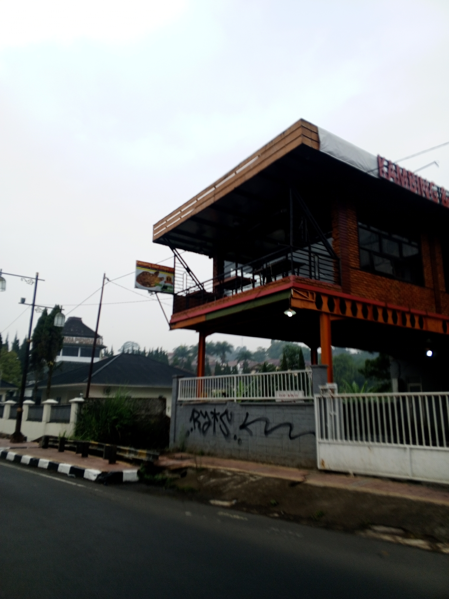 The image depicts a two-story building with an overhanging roof, constructed primarily of brick and wood. The building appears to be a restaurant or cafe, as indicated by a sign displaying food items. The setting seems to be a street in Dambulla, Sri Lanka, characterized by a paved road, sidewalks, and surrounding greenery. A white fence with vertical bars encloses part of the property, and graffiti is visible on a wall adjacent to the fence. The sky is overcast, suggesting a cloudy day.