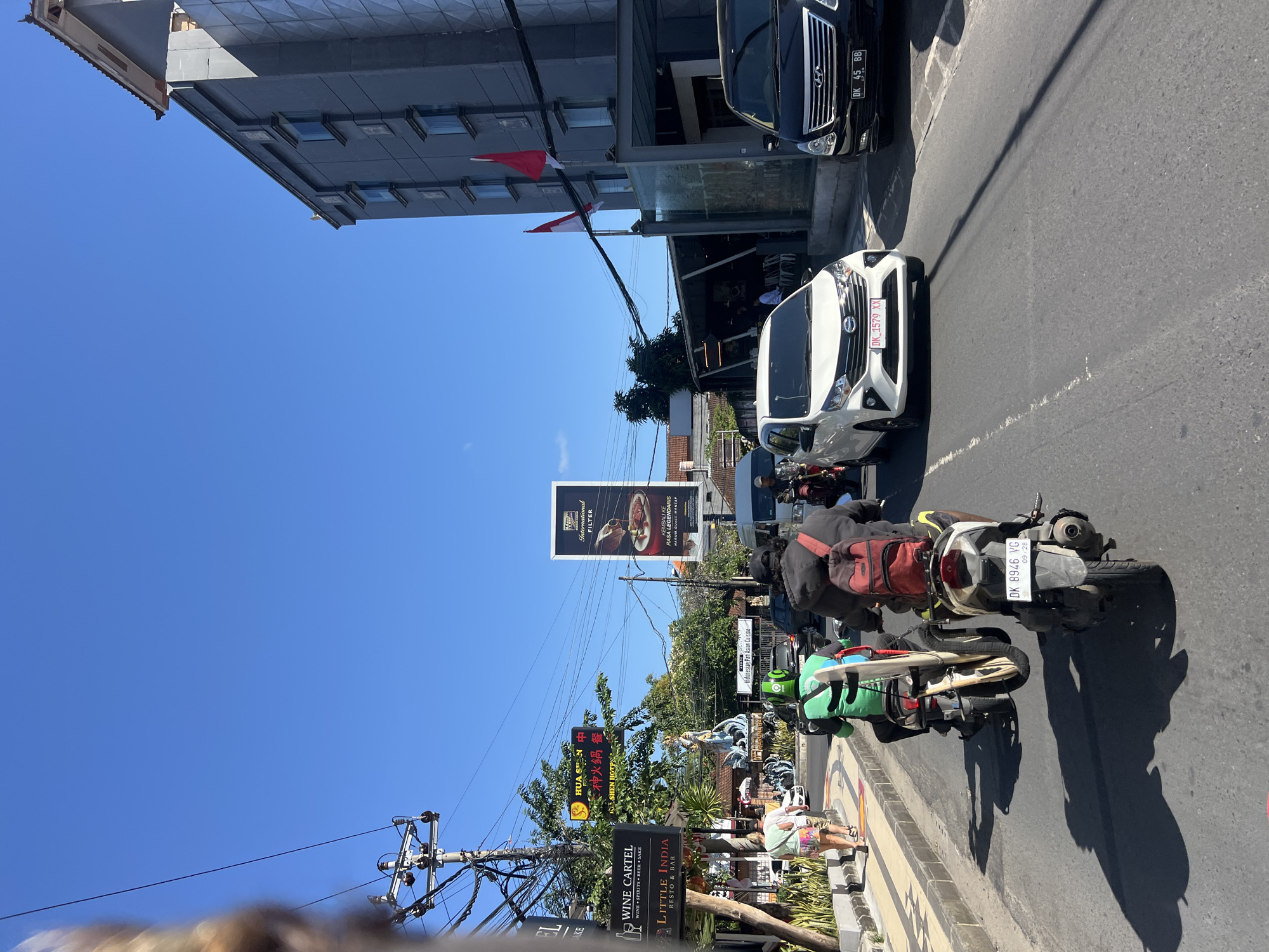 In Kuta, Indonesia, on a clear day, traffic moves along a street. A white sedan with a license plate reading "DK 1579 XX" drives in the right lane. Beside it, a black van with the license plate "DK 45 BB" also proceeds down the road. Several motorbikes are present. One motorbike in the foreground has a red backpack strapped to its rear, and another motorbike further ahead has a green backpack and a surfboard attached. People are seen walking on the sidewalk on the left, including two women. Buildings line the street, including a multi-story grey building with windows and flags on its facade. Signs for "WINE CARTEL" and "LITTLE INDIA RESTO & BAR" are visible on the left. A large billboard advertising "International Filter" is positioned in the distance. The sky is a vibrant blue, and shadows stretch across the asphalt road, indicating bright sunlight.
