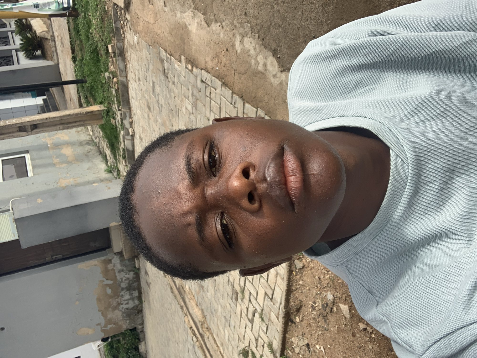 The image is a close-up, waist-up selfie taken outdoors in Jos, Nigeria. The subject, a young Black person, is wearing a light blue, textured crewneck shirt. They are positioned centrally in the frame, facing the camera with a neutral expression. The background shows an urban environment with a paved walkway, a grey, weathered building with a wooden door, and patches of grass and dirt. The lighting suggests it is daytime.