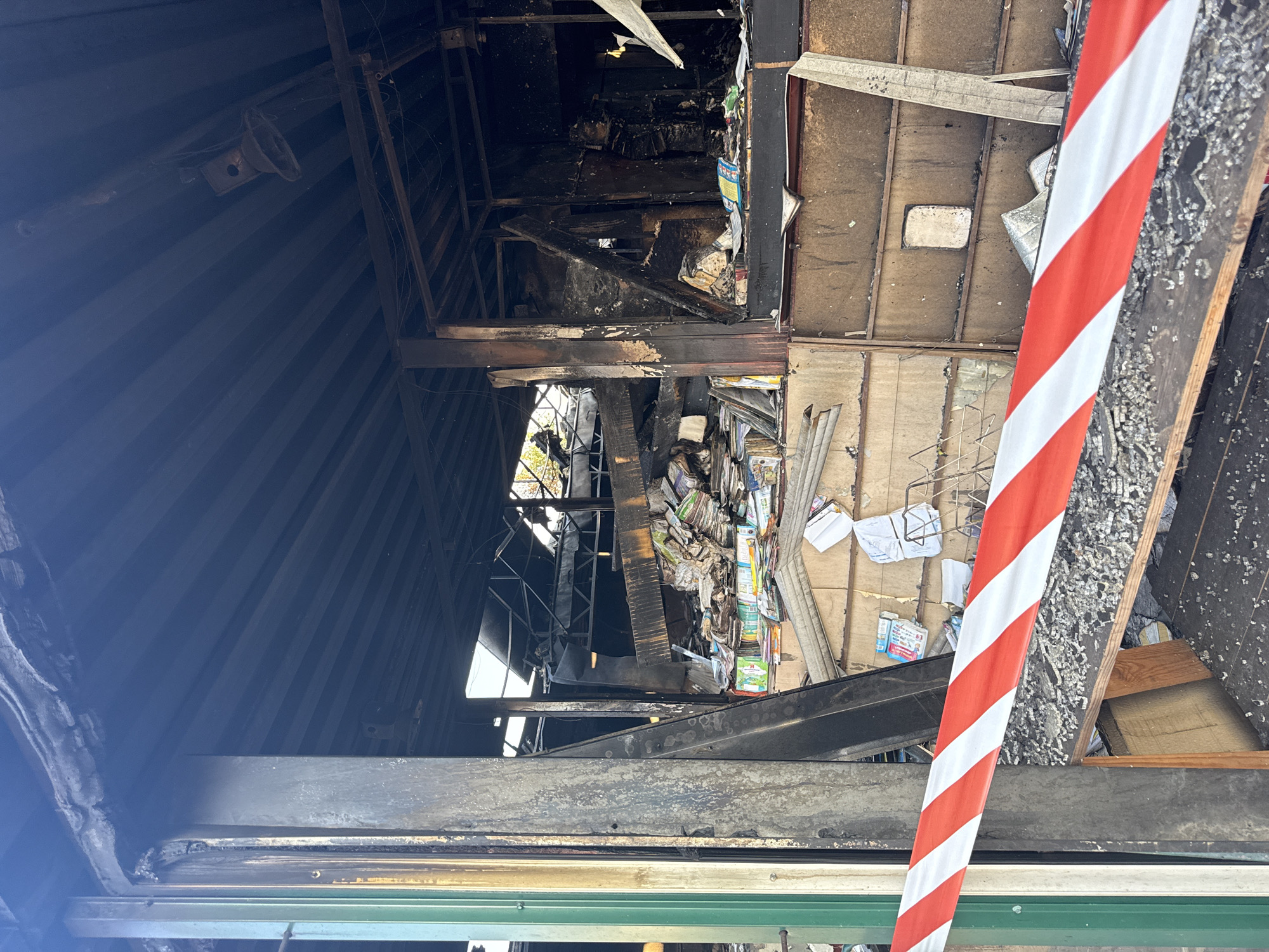 The image depicts the aftermath of a fire in a building located in Kyiv, Ukraine. The scene shows extensive structural damage, with charred beams, blackened walls, and a collapsed roof. Piles of debris, including what appear to be books and papers, are scattered throughout the interior. A red and white striped caution tape is visible in the foreground, indicating a restricted area. The environment suggests a commercial or public space that has been severely impacted by the fire.