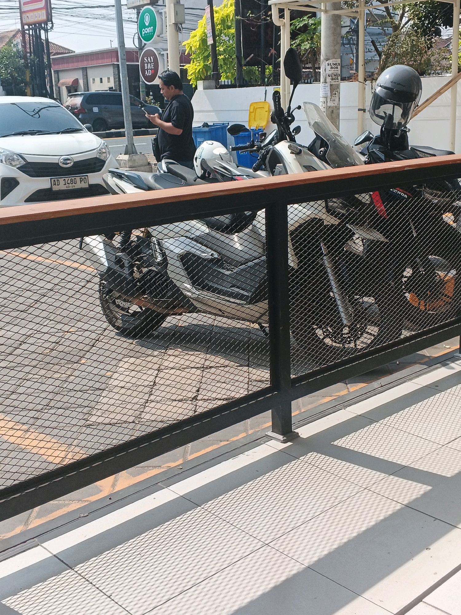 In the city of Genuk Barat, Indonesia, on a sunny day, a man in a black t-shirt is standing next to parked motorcycles, looking at his phone. He is wearing a dark shorts. In the background, there is a white car with the license plate "AD 1480 PO". A street sign for "POINT" and "SAY BRE AD" is visible. Several motorcycles are parked, one with a gray helmet on it. A black metal fence with a wooden railing runs across the foreground, casting a shadow on the tiled ground. The scene appears to be a street or a parking area, with buildings and trees in the background.