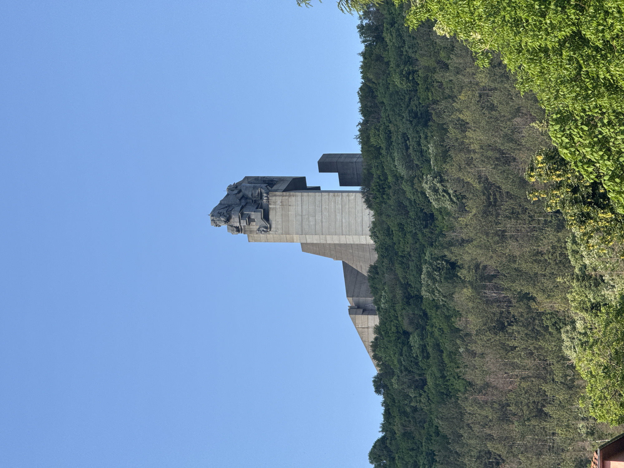 The image shows the Founders of the Bulgarian State Monument in Shumen, Bulgaria. A massive concrete structure with a sculptural group depicting historical figures stands against a clear blue sky. The monument is situated on a hill covered with dense green vegetation and trees.