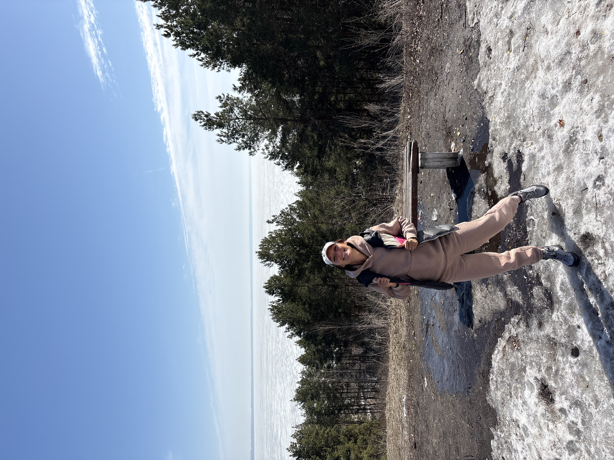 A woman sits on a bench overlooking a body of water on a partly sunny day.
The woman is wearing a tan-colored sweatsuit, a dark puffy vest, and a white cap. She smiles towards the camera.
The body of water, possibly the Volga River, stretches to the horizon. Trees line the shore and there are patches of snow on the ground. The sky is blue with streaks of clouds.
The scene may be taking place in the city of Ulyanovsk, Russia.