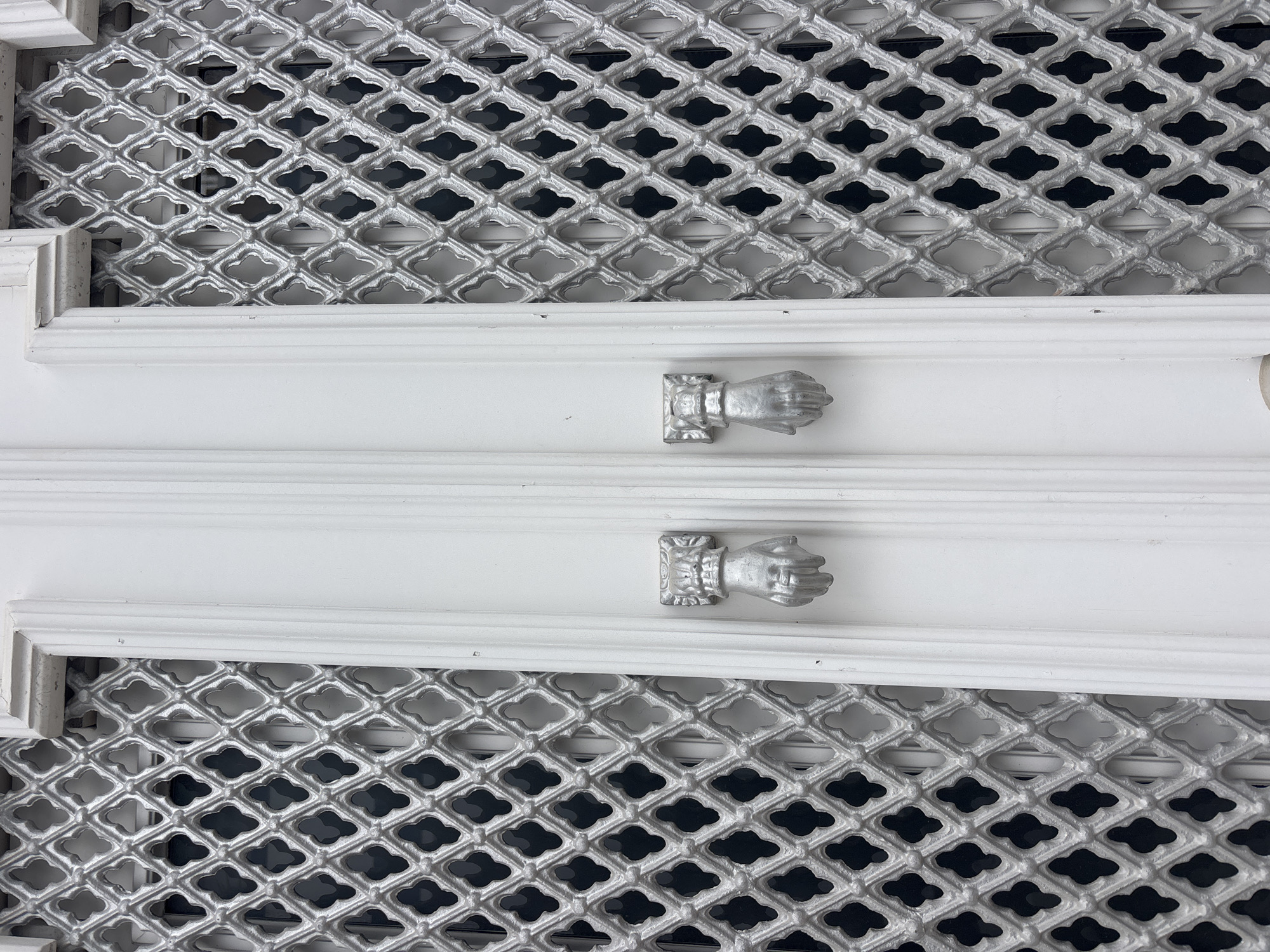The image shows a close-up of a white door with two decorative hand-shaped door knockers. The door is flanked by ornate silver metal grilles. The grilles have a repeating diamond pattern with smaller diamond-shaped cutouts. The door itself is also white and has vertical fluting in the center. The door knockers are in the shape of a clenched fist with the knuckles facing outward, mounted on a rectangular base. The scene appears to be an entrance to a building, possibly a home or a shop, in the city of Tavira, Portugal. The lighting suggests it is daytime, with some shadows cast by the grilles.