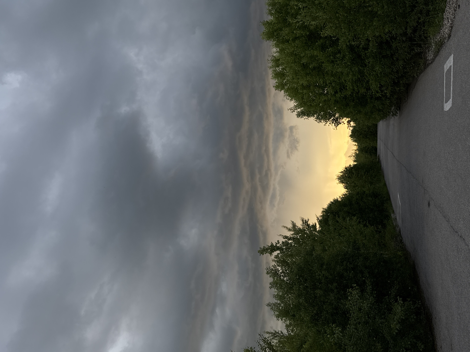 An empty asphalt road leads into a distance, bordered on both sides by lush green trees. The sky above is filled with dark, ominous clouds, with a sliver of golden light breaking through on the horizon. This scene captures a moment of impending weather change, with the road acting as a pathway into the unknown. The location is the city of Perm, Russia.