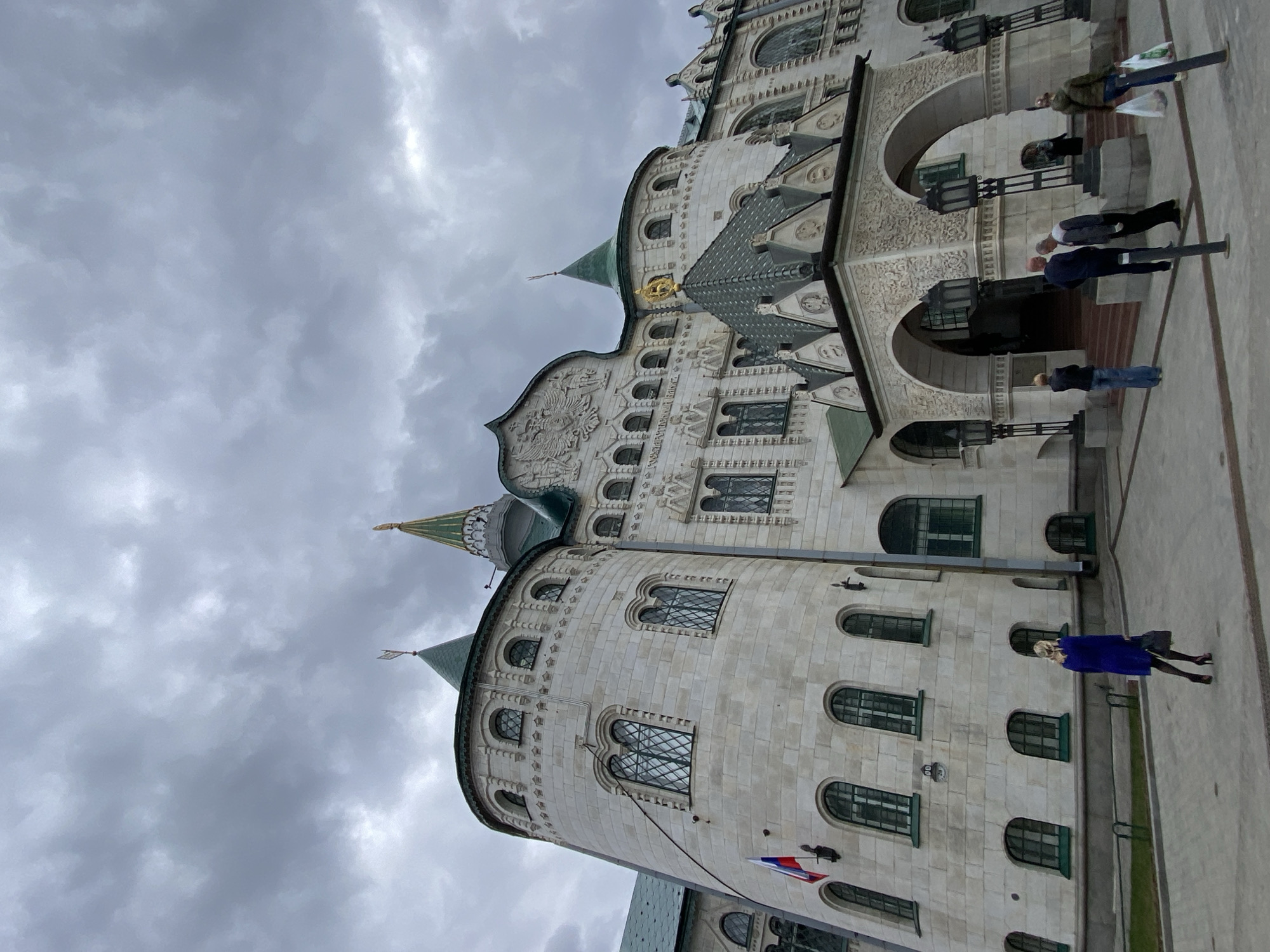 The image displays a grand, multi-storied building under a heavily overcast sky, with numerous grey clouds. The building is constructed from light-colored stone blocks and features intricate architectural details, including decorative carvings, patterns, and various arched windows, some fitted with grilles. Several green, pointed spires or small domes protrude from the roofline.
On a prominent section of the roof, a golden, ornate relief, resembling a double-headed eagle, is visible. Below, the facade includes two large arched entrances, flanked by decorative lampposts.
Several people are present in front of the building on a paved ground. On the left side, a woman in a bright blue coat and black tights/heels walks towards the right. Near the arched entrance on the right, multiple individuals are gathered, some appearing to be standing or walking. One person wears a brown jacket and carries bags.
A small Russian tricolor flag (white, blue, red) hangs from a pole attached to a cylindrical tower section of the building on the left. Visible Cyrillic text carved into the building's facade, specifically above the central entrance, reads "ГОСУДАРСТВЕННЫЙ БАНКЪ" (State Bank). The scene depicts a historical or official building in Nizhny Novgorod, Russia, on a cloudy day.