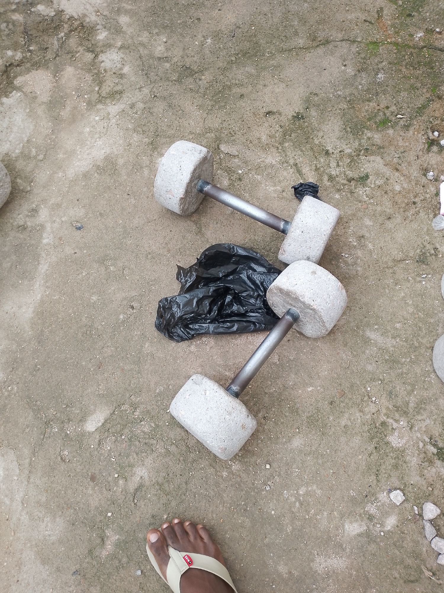 In an outdoor scene on an unpaved surface, two homemade dumbbells are visible, resting on the ground. Each dumbbell consists of a cylindrical concrete weight attached to a metal bar. One dumbbell is positioned diagonally from the upper left to the lower right, while the other is placed above and to the left of it, oriented horizontally. A crumpled black plastic bag lies between the dumbbells. In the lower left corner of the frame, a person's foot is visible, wearing a light-colored sandal. The ground is a mixture of dirt and small stones, with patches of moss.