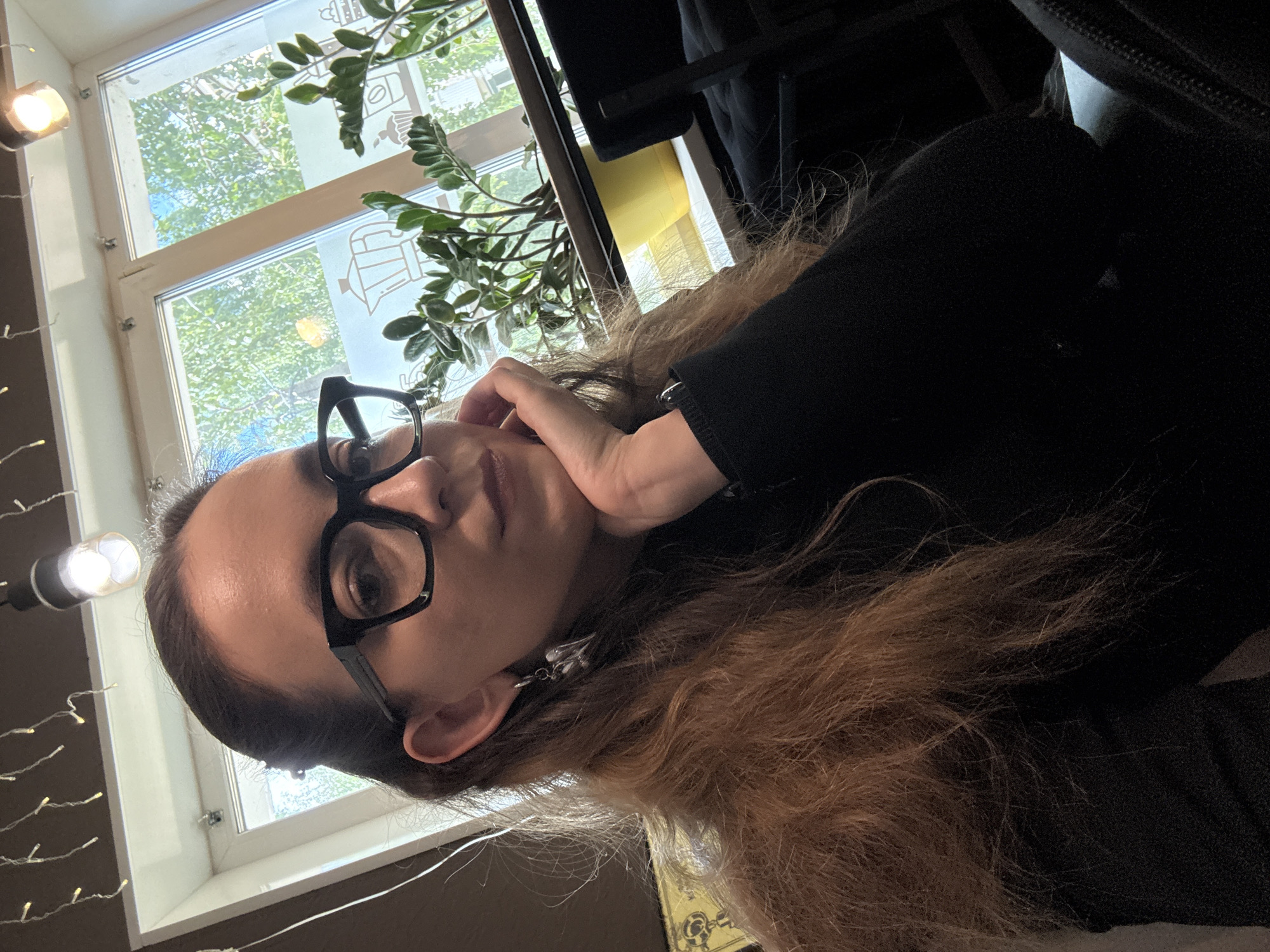 A woman with long, brown hair and black-rimmed glasses is taking a selfie. She is wearing a black top and a dangling silver earring shaped like wings. Her right hand is resting on her cheek. The background shows a window with a view of green trees. There is a string of fairy lights hanging from the ceiling and a plant in a yellow pot on the windowsill. The setting appears to be a cafe in Chaikovsky, Russia, as indicated by the presence of a moka pot illustration on the window and what looks like typical cafe seating.