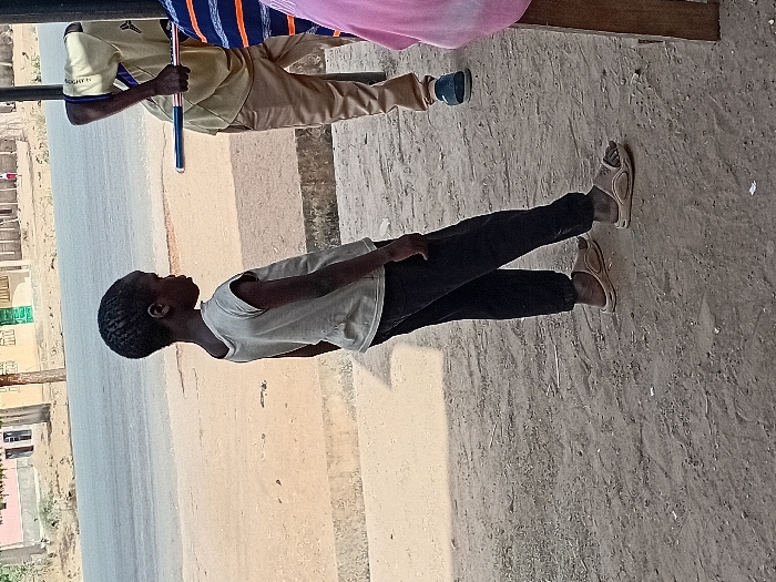 The image depicts a daytime scene in Dabawa, Nigeria, featuring a young child and partially visible adults on a dirt ground adjacent to a paved road. In the foreground left, a child with dark skin and short, dark hair wears a light-colored sleeveless t-shirt, dark long pants, and light brown open-toed sandals. The child is positioned facing slightly right, with head turned further right, observing something outside the frame. To the child's right, an adult male, partially visible and bent over, wears a light-colored short-sleeved collared shirt, light brown pants, and dark blue closed-toe shoes. This adult holds a long, slender object with blue, white, and red sections. Another individual, partially visible to the right, wears a blue and orange striped shirt. Further right, a section of pink fabric and a wooden structure are present. The immediate ground is dirt, transitioning to a concrete curb, and then a gray asphalt road. Across the road, multiple single-story buildings, some light-colored and one with a prominent green door, are visible under bright lighting conditions.