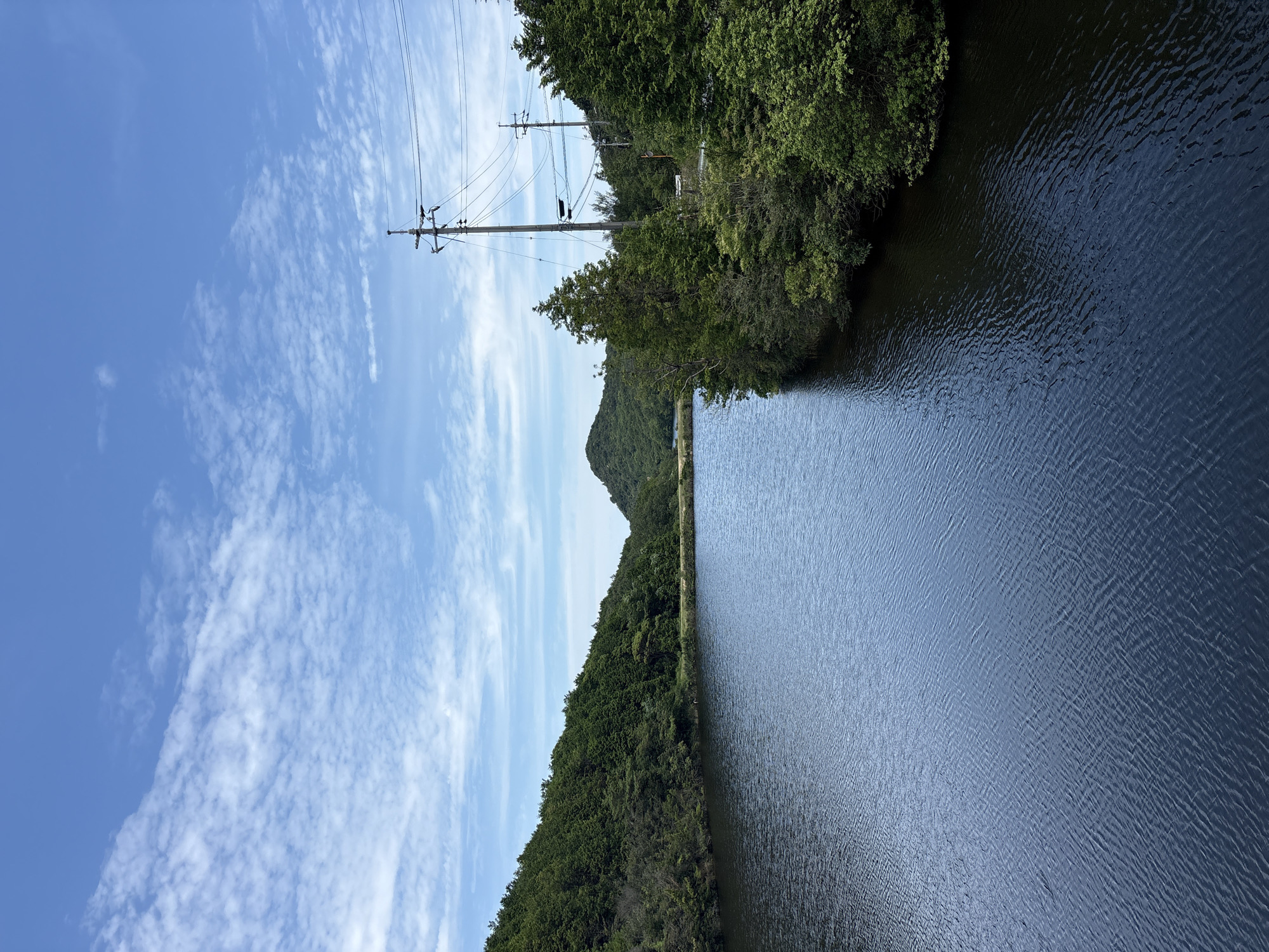 The image shows a serene landscape with a body of water in the foreground, flanked by lush green hills and trees. The water is dark blue with subtle ripples, reflecting the blue sky above, which is dotted with scattered white clouds. On the right side of the water, a line of trees and vegetation meets the water's edge, and beyond that, there are utility poles with electrical wires stretching across the scene. The background features rolling hills covered in dense green foliage, with a faint misty atmosphere in the distance. The overall impression is one of natural beauty and tranquility, possibly a reservoir or a large pond within a hilly, forested area. This scene is located in the city of Naoshima, Japan, a place known for its art islands and natural landscapes.