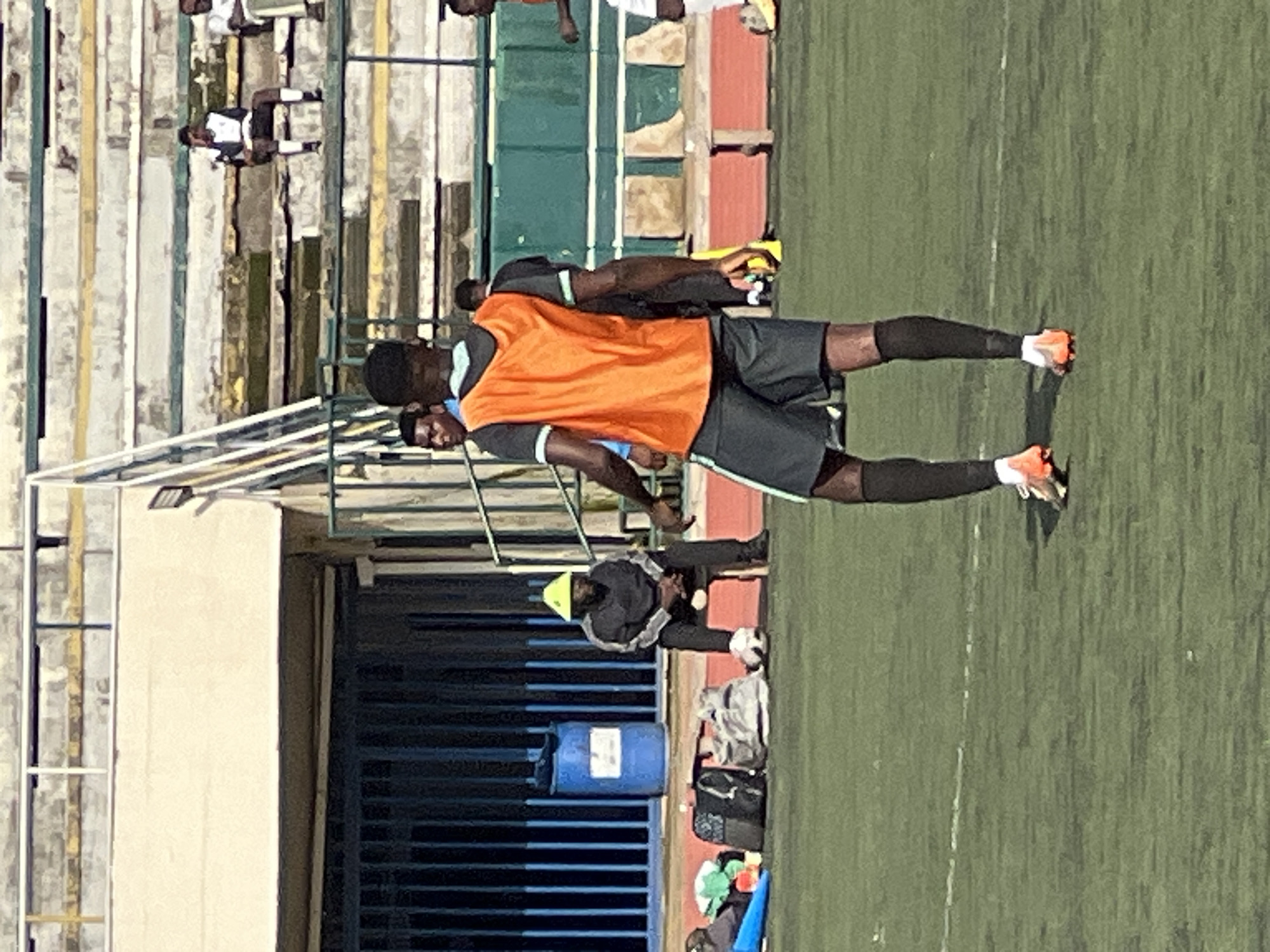 In the city of New Stadium, Nigeria, on a sunny day, several individuals are present on a green turf field, likely a sports arena. In the foreground, a young Black male wearing an orange training bib over a dark t-shirt and grey shorts is standing, facing away from the camera, with his back to the stands. He is wearing black socks and orange and white soccer cleats. To his left, another Black male is partially visible, looking towards the person in the orange bib.
In the mid-ground, to the left, a Black male is seated on a dark chair, wearing a grey tracksuit and a bright yellow conical hat. Next to him are several sports bags and a blue barrel. Behind them is a metal fence.
In the background, rows of stadium seating are visible, with a few individuals seated in them, observing the scene on the field. The overall environment suggests a sports training or practice session.