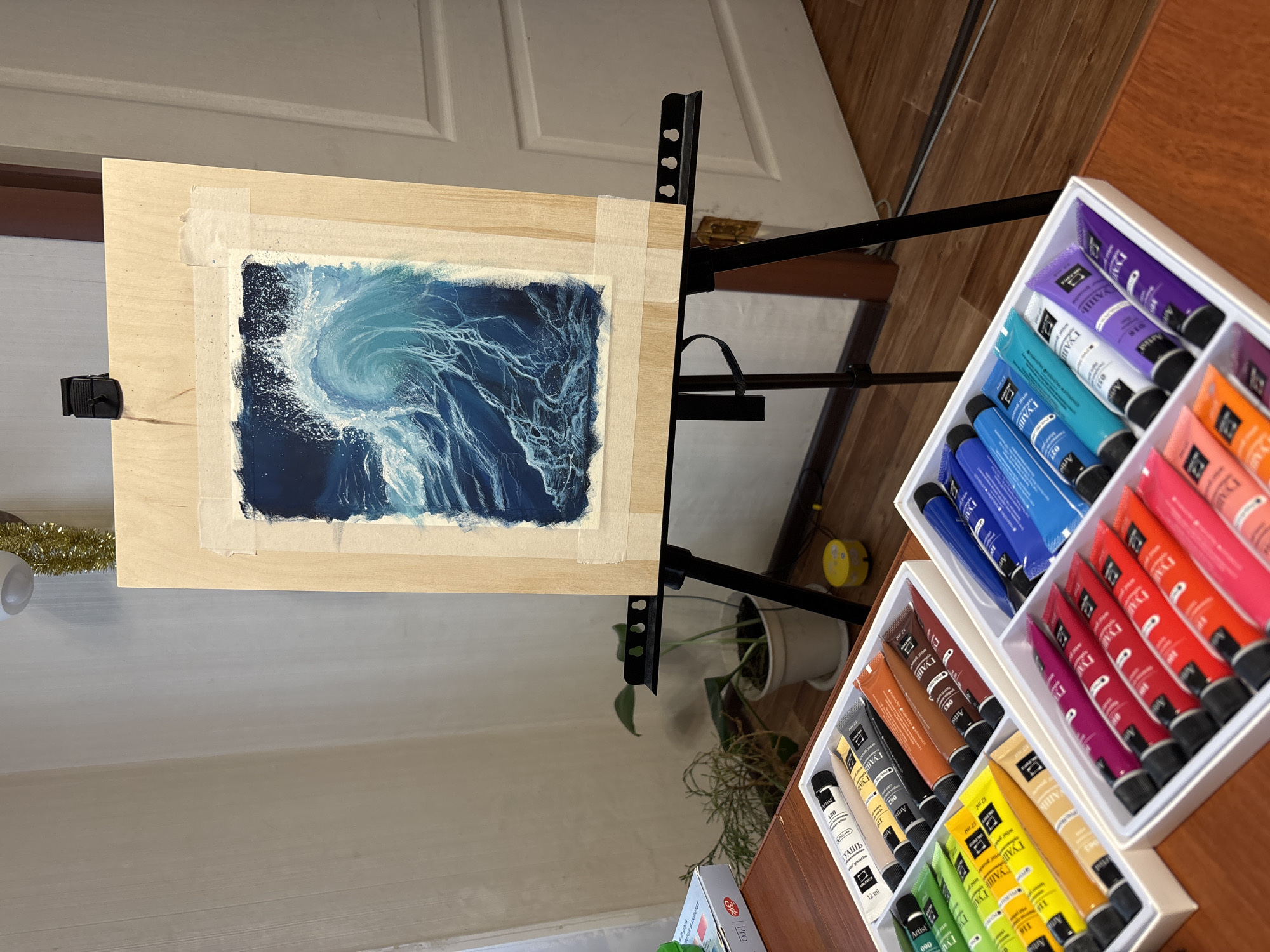 Here is a detailed description of the image:
**General Scene:**
The image shows an artist's workspace. A painting of a wave is mounted on an easel. To the side, there are two sets of gouache paints organized in white containers.
**Subjects and Objects:**
* **Painting:** The central focus is a painting depicting a large, swirling wave. It uses a palette of blues, whites, and some dark shades to create depth and movement.
* **Easel:** The painting is held on a black easel, typical of art studios.
* **Paints:** Two sets of gouache paints in white containers are visible, filled with tubes in a spectrum of colors (purples, blues, oranges, reds, browns, yellows, and greens). Each container appears to hold a different range of hues.
* **Background:** The background consists of a light-colored wall, part of a white door with panel details, and a wooden surface (table or floor).
* **Other Objects:** Other objects are partially visible, including a yellow object (likely a container), a potted plant, and gold tinsel.
**Actions/Interactions:**
* The scene suggests the artist is in the process of creating or has recently completed the wave painting.
**Environment:**
* **Indoor Setting:** This is an indoor workspace, possibly a home studio, indicated by the domestic furnishings and the organized arrangement of art supplies.
* **City and Location:** No information in the image indicates that this scene is in the city of Izhevsk, Russia.