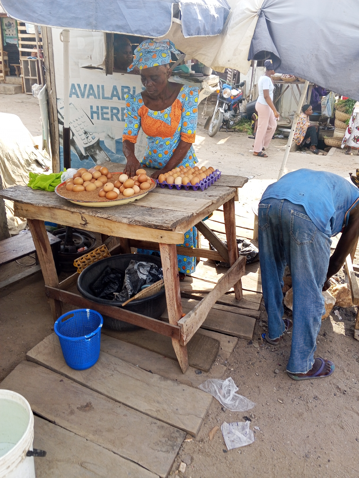 A woman in a patterned blue and orange dress and headscarf stands behind a wooden table laden with eggs in Zango-ndaji, Nigeria. Some eggs are piled on a metal platter, while others are in a purple carton. A man in a blue shirt and jeans bends over near the table. Underneath the table is a black bucket with refuse, and a blue plastic bucket sits nearby on wooden planks. Debris litters the ground. An umbrella provides shade. Other people and a motorcycle are visible in the background.