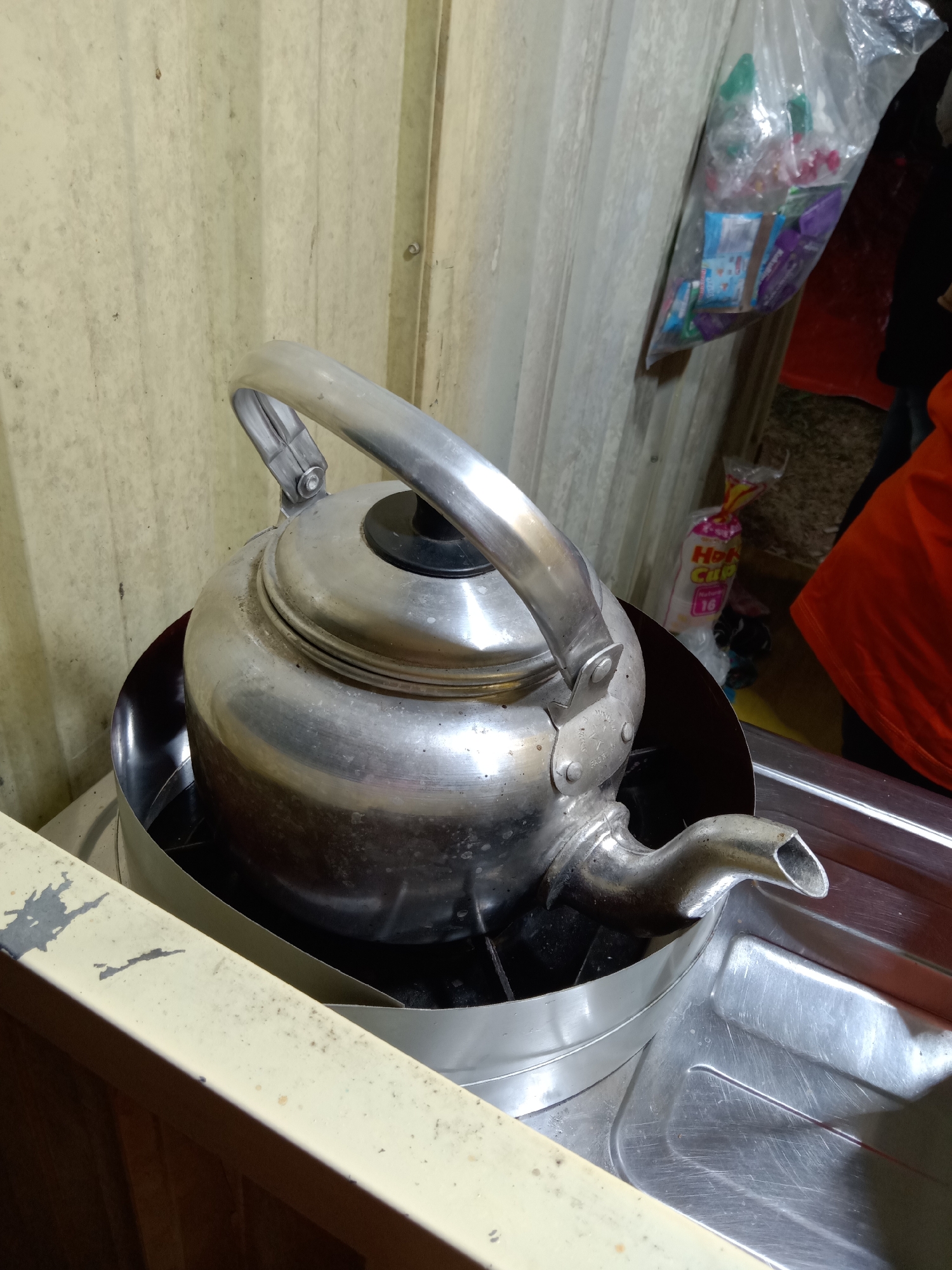 The image depicts a metal teapot placed on a stove in an indoor setting. The teapot is silver and has a black lid. It is positioned on what appears to be a portable gas stove, with a metal wind guard around its base. The stove is situated on a metallic surface that looks like part of a kitchen counter. In the background, a light-colored, corrugated wall is visible. To the right, a clear plastic bag filled with various packaged items, possibly snacks or instant food, is hanging. Partially visible behind the bag is a person wearing an orange garment. The scene suggests a simple, utilitarian kitchen environment. The image is taken from a slightly elevated angle, looking down at the teapot and stove. The lighting is somewhat dim, with the focus on the teapot and its immediate surroundings. There are no visible indications of the location being specifically Stiaji, Indonesia, within the image itself.