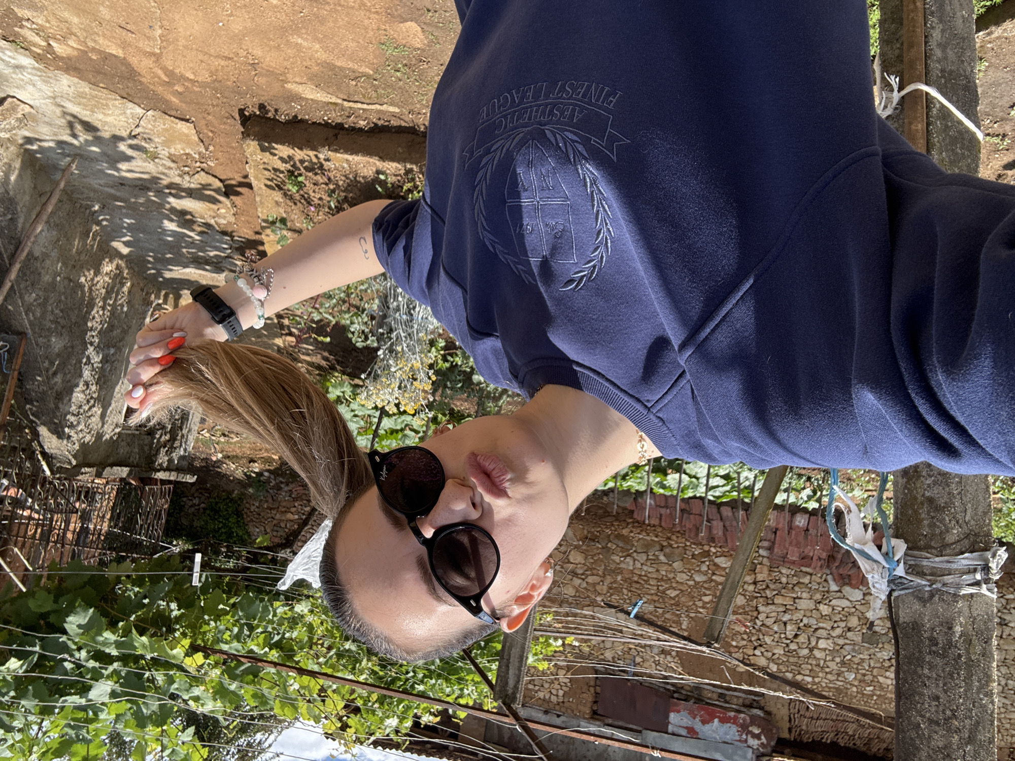 A young woman with blonde hair styled in pigtails, wearing a dark blue sweatshirt and black sunglasses, is taking a selfie outdoors. She is leaning her head to the side and puckering her lips. The background shows a rustic, outdoor setting with a stone wall, a wooden pole, and vegetation. This scene is consistent with the rural outskirts of Prolom, Bulgaria, characterized by traditional stone buildings and natural landscapes.