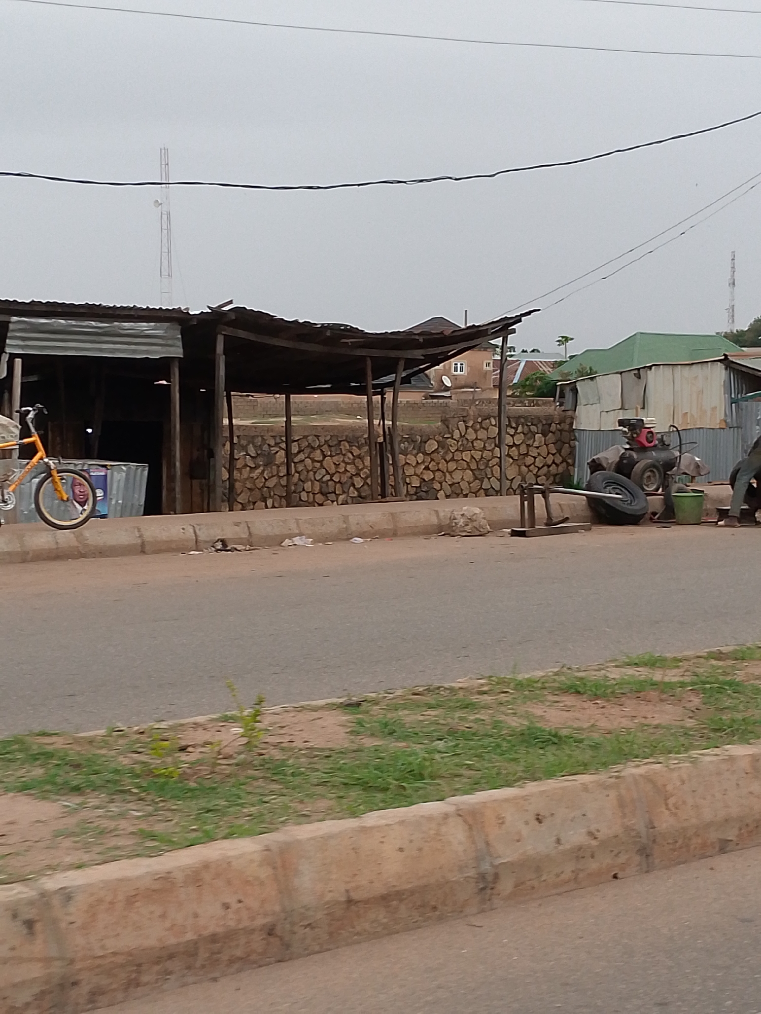 The image depicts a street scene in Bukuru, Nigeria, on an overcast day. The foreground shows a street with a concrete curb separating it from a strip of grass. Behind the curb, a road with little traffic extends towards the background. Along the roadside, there are several makeshift structures, including stalls and shops made from wood and corrugated metal. One structure on the left is partially obscured by an orange bicycle leaning against it. To the right, a stone wall serves as a backdrop for a tire repair operation, indicated by an air compressor, tires, and repair tools. A person can be seen working nearby, but only their legs are visible. In the background are several buildings, including houses and businesses with corrugated metal roofs. Power lines stretch across the sky, and a cell phone tower is visible in the distance.
