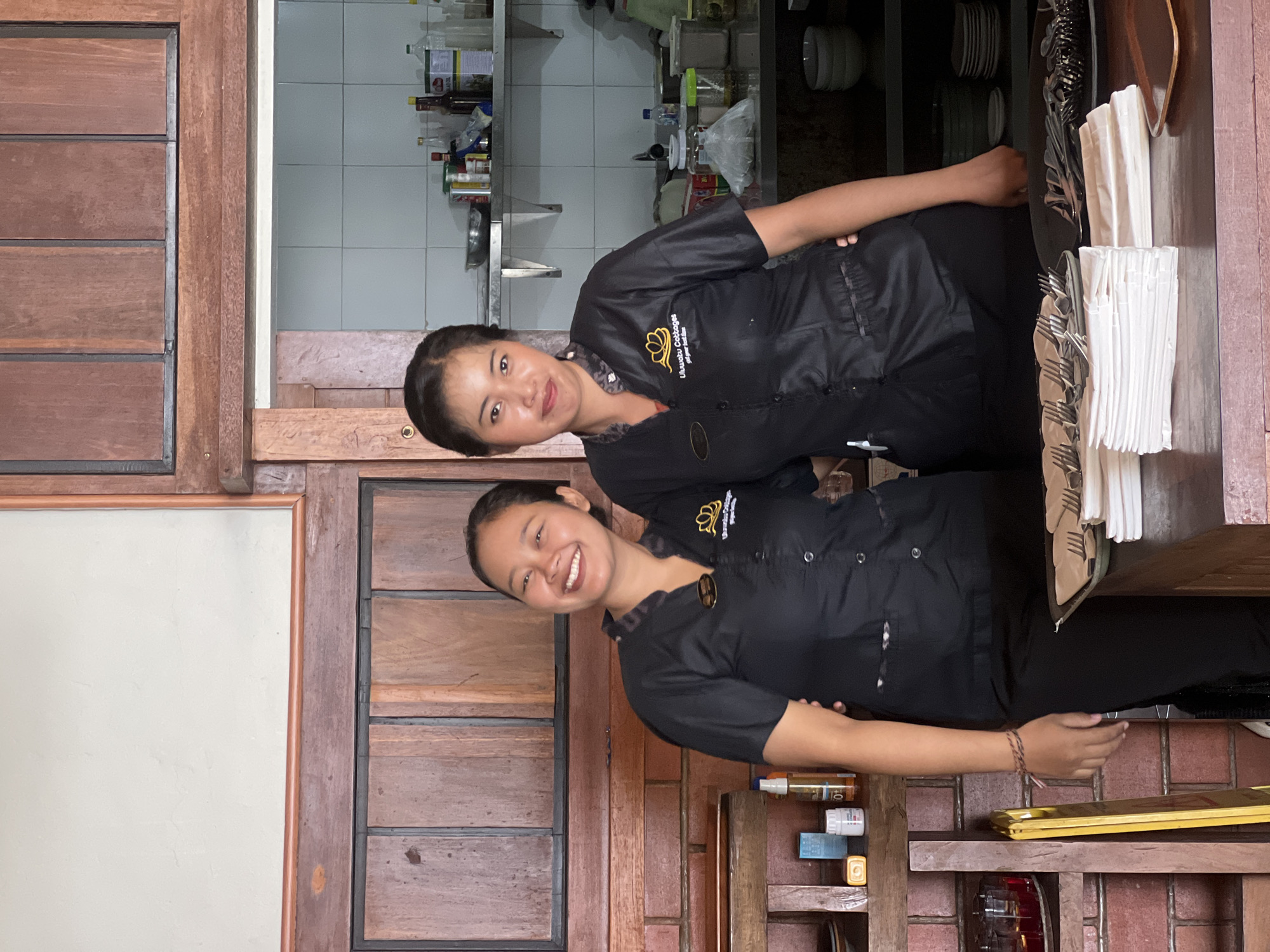 Two women stand in front of a doorway with exposed brick and wood paneling at a business called Uluwatu Cabbages in Kuta Selatan, Indonesia. The women are wearing black uniforms with a logo. The woman closest to the viewer is smiling. Silverware and dishware can be seen in the background.