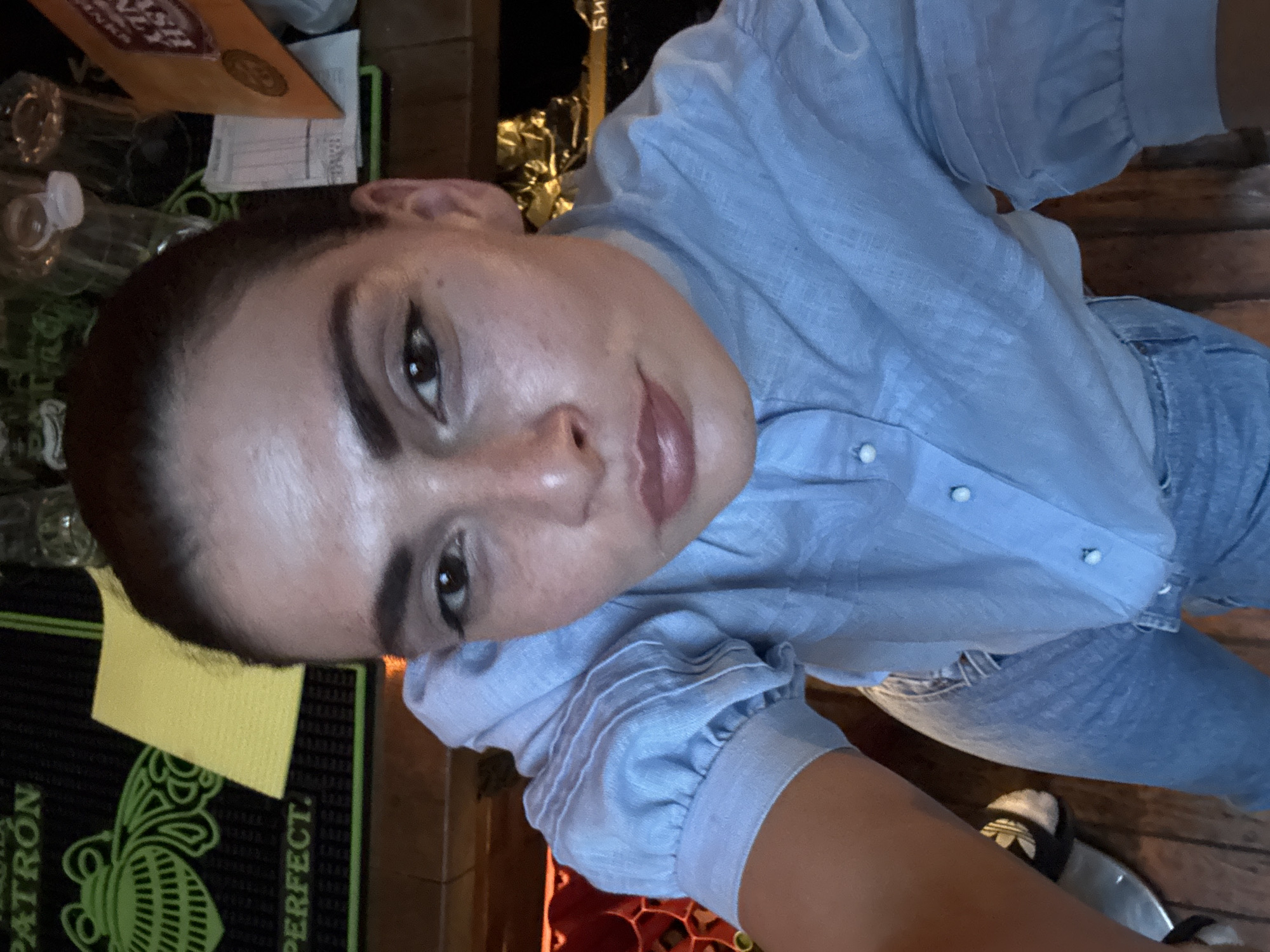 This angled selfie features a young woman looking directly at the camera, likely taken indoors in Sofia, Bulgaria. She has dark hair pulled back, dark eyes, and a neutral to slightly smiling expression with full lips. She is wearing a light blue, collared button-up shirt with distinctively gathered or puffed sleeves and small white or pearl-like buttons. Her lower half is clad in light blue denim jeans, and black sandals, possibly Adidas slides, are partially visible on her feet.
The background suggests a casual indoor setting, likely a bar or a counter area. To the left, several clear plastic bottles are visible on a surface, alongside a bright yellow napkin or cloth. A prominent black bar mat with a green, intricate design, resembling a beetle or insect, displays the text "PATRON" and "PERFECT." Further in the background, out-of-focus elements include an orange envelope or folder, wooden textures, and crumpled metallic material. The lighting appears to be ambient indoor light.