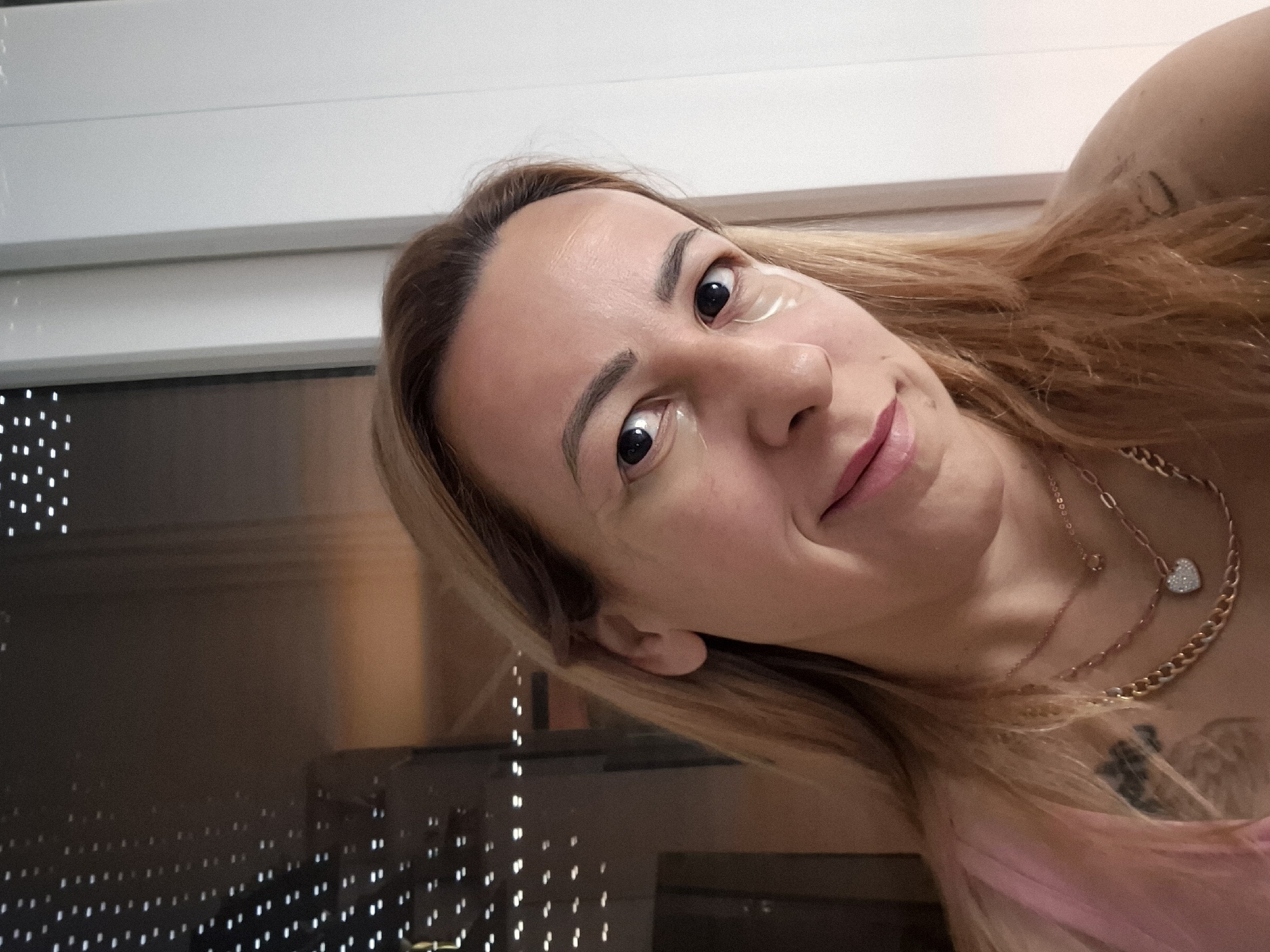 A woman with blonde hair and visible under-eye patches smiles for a selfie. She is wearing a pink top and multiple necklaces, including a gold chain and a rose gold chain with a heart-shaped pendant. A tattoo is visible on her left shoulder. The background appears to be indoors, with a window showing a blurred view of lights. The image is taken from a close-up perspective.