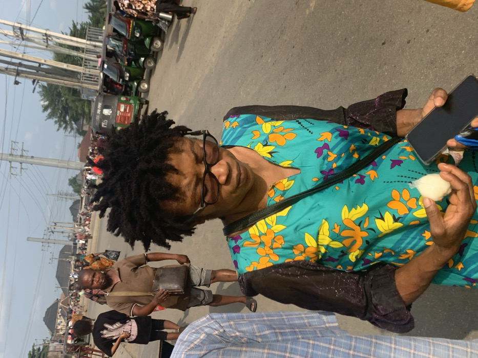 This image depicts an outdoor urban scene in Ura Nta Umuarandu, Nigeria, during daylight hours. In the foreground, a female subject with dark dreadlocks and dark-rimmed glasses wears a teal top adorned with orange, yellow, and purple floral patterns, featuring dark, long sleeves. She holds a small, white, plastic-wrapped spherical object in her left hand and a black smartphone in her right, with her head oriented downwards. To her immediate left, a male is partially visible, wearing a light blue and white checkered shirt.
In the midground, a male with short dark hair, dressed in a brown polo shirt, grey shorts, and dark flip-flops, carries a dark leather messenger bag across his left shoulder and looks forward. Several other pedestrians are scattered throughout the midground and background, indicating a populated area. The background features an asphalt road where a line of green and red auto-rickshaws, identified as "Keke Napep," are parked on the right side. Overhead, numerous utility poles and an intricate network of electrical wires are visible against a bright sky. Distant buildings with light-colored roofs complete the urban landscape.