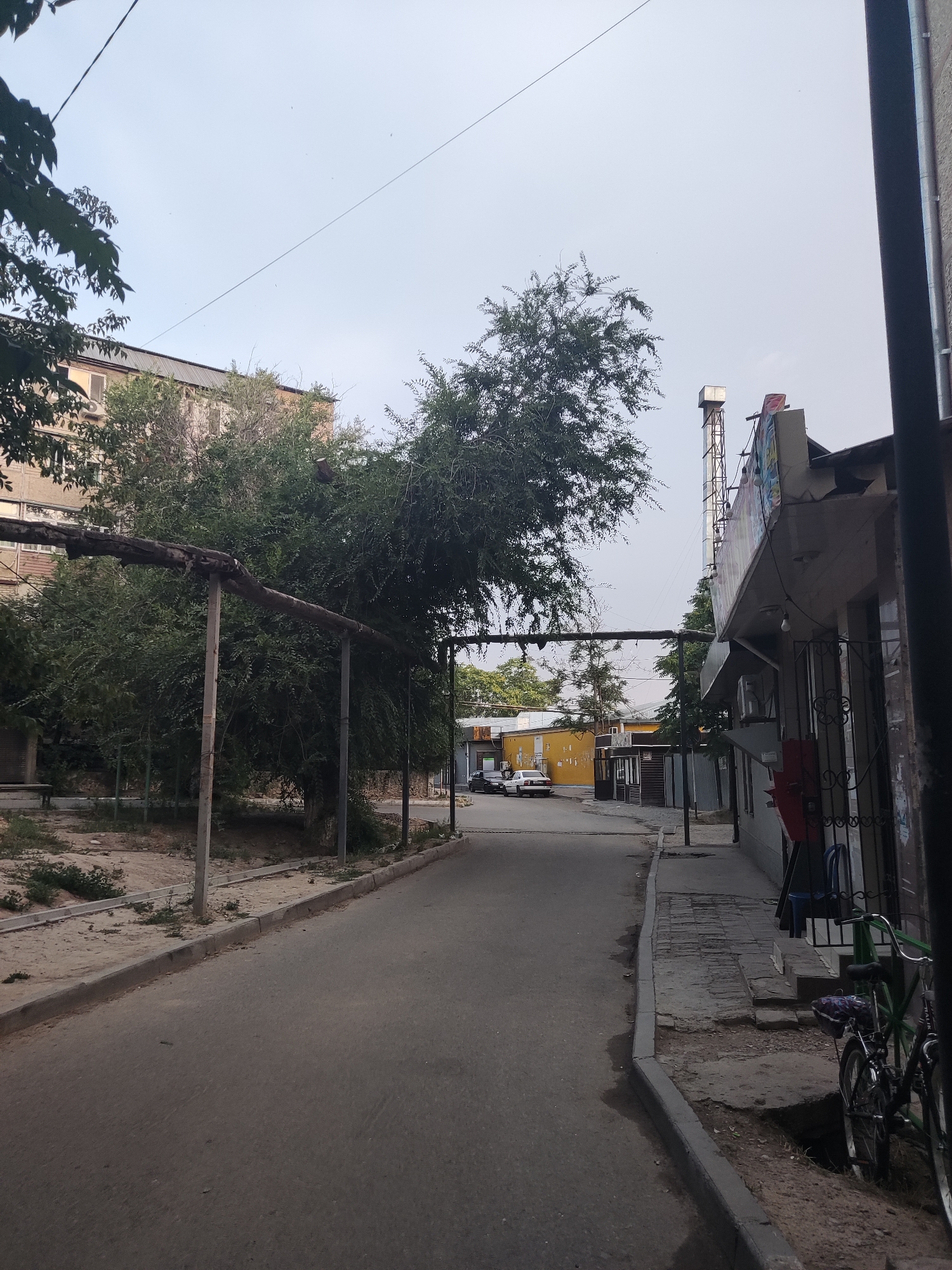 The image depicts a street scene in Shymkent, Kazakhstan. On the left side of the paved road, a sidewalk area with a dirt and gravel surface is visible, lined with a concrete curb. A large, leafy tree dominates this side, with a thick, weathered pipe supported by metal posts running horizontally in front of it. Behind the tree, a light-colored apartment building with visible windows and an air conditioning unit can be seen.
The right side of the road features a sidewalk with uneven paving stones and a curb. Adjacent to the sidewalk is a building with a light-colored facade, a dark pipe running vertically along its side, and a patterned metal gate. A red box is mounted on the wall near the gate. Further down the sidewalk, a bicycle with green handlebars is parked.
The road extends into the distance, where more buildings are visible. To the left, a yellow building and a smaller structure with a dark corrugated metal fence are present. Two cars, one white and one dark, are parked on the street in front of these buildings.
In the distance, a tall, narrow structure, possibly a ventilation shaft or industrial pipe, rises above the buildings. The sky above is overcast, with a thin power line stretching across it. The overall impression is of a somewhat utilitarian urban environment, possibly a back street or an industrial-adjacent area.