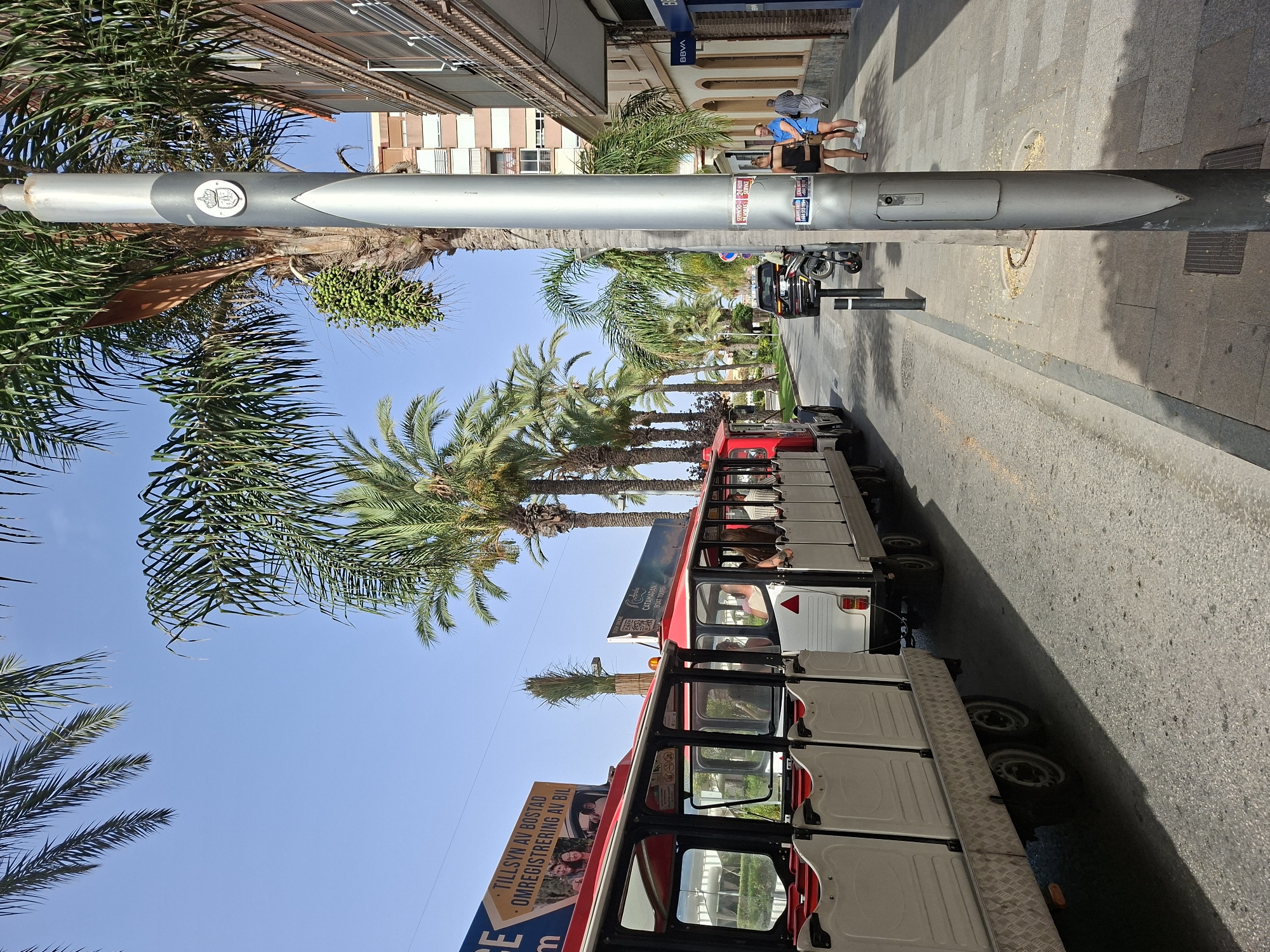 The image depicts a sunny street scene in Torrevieja, Spain, likely a popular tourist area due to the presence of a red and white open-air tourist train. The train, with several carriages and passengers visible, is parked on the right side of the road, casting a shadow. Palm trees line the street, indicating a warm climate. On the sidewalk to the right, two women are standing near a silver lamppost that has several posters attached to it. A black car and a motorcycle are parked further down the street, and a building with shops, including one with a "BBVA" sign, is visible in the background. The sky is clear blue.