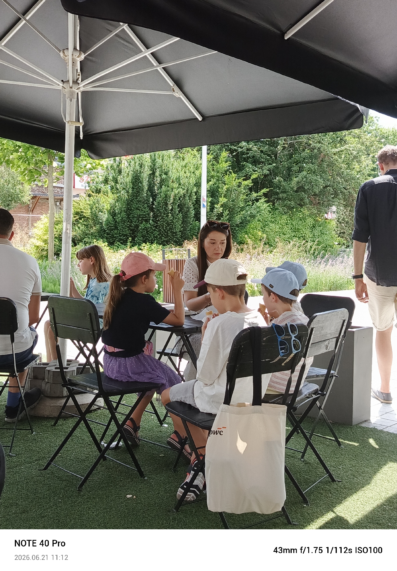 A group of people, including children and adults, are seated at tables outdoors, seemingly at a cafe. A large black umbrella shades the area. One child in a pink hat is eating something, while another child in a white shirt and cap is also holding food. A woman in a white top is seated at the table. To the right, a man in a dark shirt and light shorts stands, looking away from the camera. The environment appears to be a paved area covered in artificial turf, with lush green trees and foliage in the background. The scene is well-lit, suggesting daytime. The watermark at the bottom indicates the image was taken with a "NOTE 40 Pro" on "2026.06.21 11:12", with camera settings "43mm f/1.75 1/112s ISO100". There is no specific visual information in the image to confirm it is located in Piaseczno, Poland.