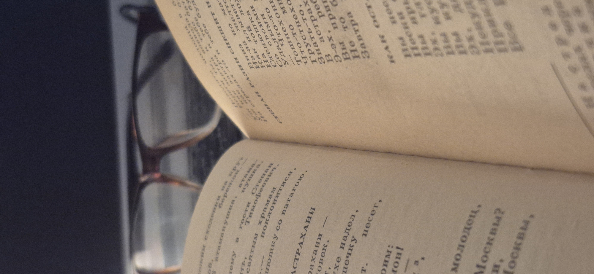 An open book with aged, yellowish pages lies on a surface, viewed from an elevated, angled perspective. A pair of eyeglasses rests on the crease of the open book, suggesting a pause in reading or preparation for it. The background is softly blurred, showing dark, indistinct shapes, likely part of a table or desk where the book is placed. The lighting is soft and diffuse, creating a calm atmosphere without harsh shadows.
The visible text on the pages is in Russian Cyrillic. The upper page includes phrases such as "СТЕПАН РАЗИН СПЕШИ" (Stepan Razin hurry), "Из-за той лодки" (Because of that boat), "Как плывет" (How it sails/swims), and mentions "Астрахани" (Astrakhan) and "Москвы" (Moscow). Words like "Грустнехонько" (very sadly) and "Тошнехонько" (very sickeningly) also appear. The lower page continues with words like "атаманушка" (little ataman), "Степан Тимофеевич" (Stepan Timofeevich, referring to Stepan Razin), "святым храмам" (holy temples), and "молодец" (brave fellow). The content strongly indicates that the book is a historical or folkloric work, possibly poetry or songs, centered around the legendary Cossack leader Stepan Razin and his exploits.
The image depicts a quiet, personal moment of reading or contemplation, potentially in a home or study setting in Kryvyi Rih, Ukraine.