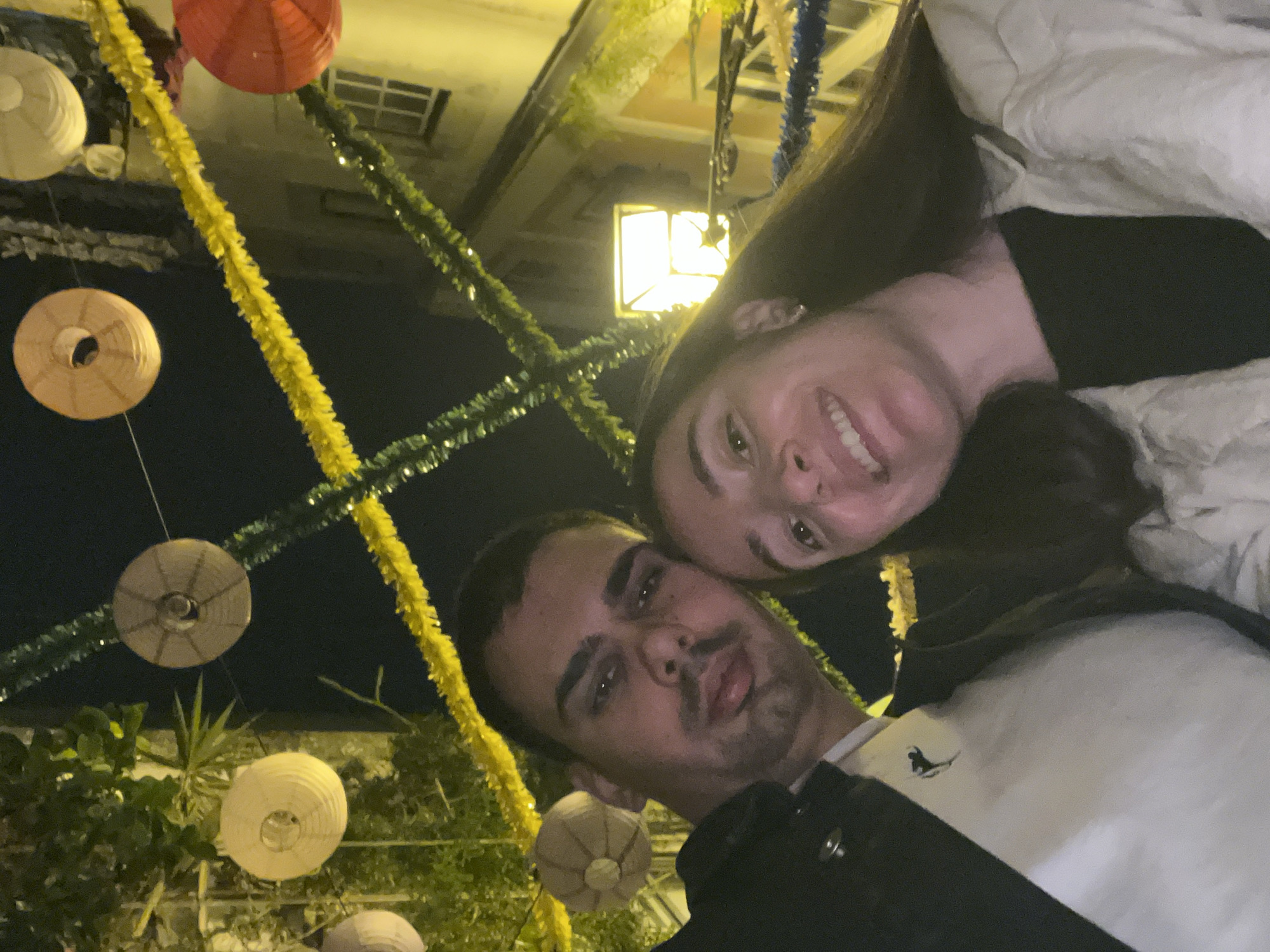 A close-up, slightly tilted selfie captures a man and a woman. The woman is positioned at the top, smiling broadly with visible teeth. The man is below her, looking at the camera with a more neutral expression.
In the background, strings of yellow, green, and blue tinsel hang alongside several spherical paper lanterns. These decorations suggest a festive or celebratory atmosphere. Behind the decorations, a building is dimly visible, featuring a window or light fixture.
The scene likely takes place in Lisbon, Portugal, during a festival or celebration where streets are adorned with colorful decorations. The lack of specific landmarks makes precise localization difficult.