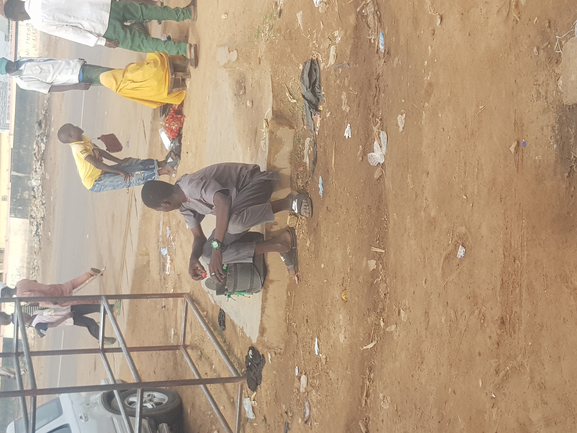 The image shows a street scene in what appears to be Kufena, Nigeria. A man is crouching near a backpack, possibly working on something with small objects. Several other people are walking along the road in the background. The surrounding area is a mix of paved and unpaved ground, with litter scattered about. A metal frame structure is in the foreground, partially obscuring a vehicle.
