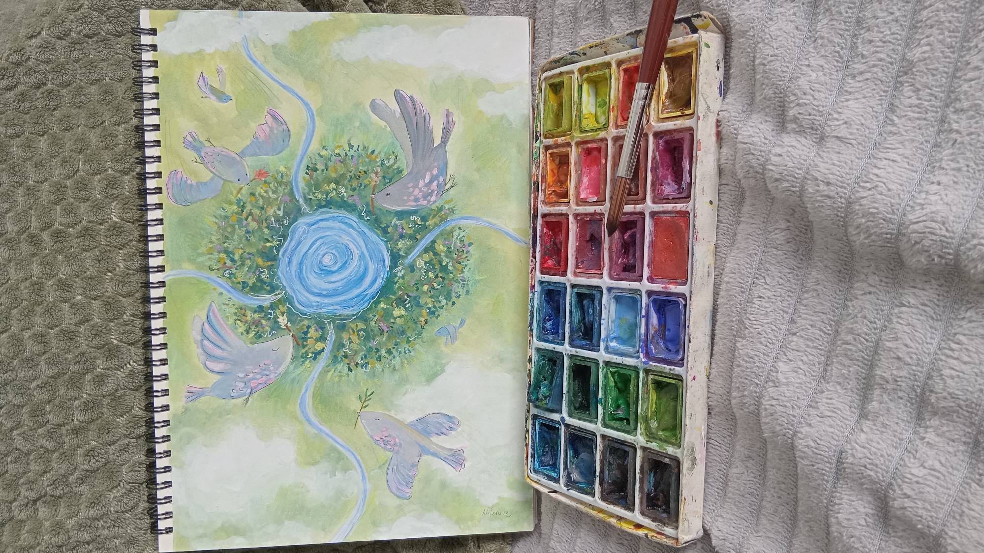 A still life photograph shows an open sketchbook with a watercolor painting next to a watercolor palette and brush, all on a textured gray surface. The sketchbook is spiral-bound and displays a painting of birds flying around a central blue swirl, seemingly within a green and white landscape. The watercolor palette contains a range of colors, with the brush currently dipped into a red-purple hue. The lighting is soft and natural. There is no indication of the location of Batumi, Georgia.