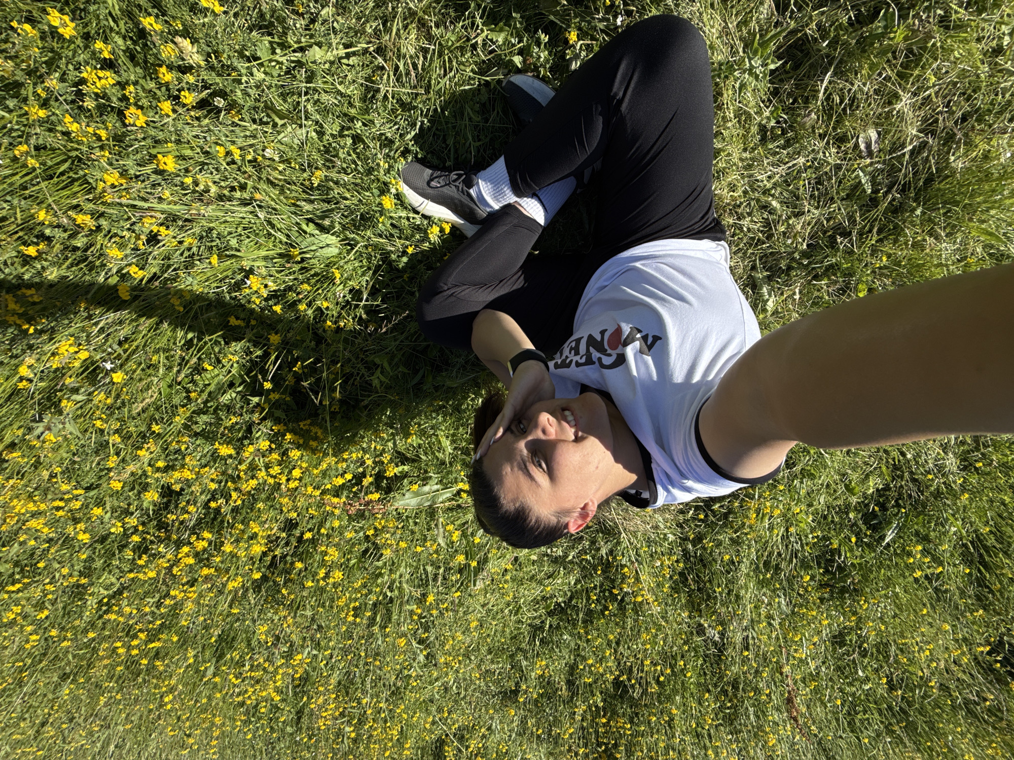 A woman is sitting in a field of yellow flowers and green grass, taking a selfie. She is wearing a white t-shirt with black trim and black leggings. She has dark hair pulled back in a ponytail. The woman is smiling and has her hand on her forehead. The image appears to have been taken on a sunny day. The location is Yundola, Bulgaria.