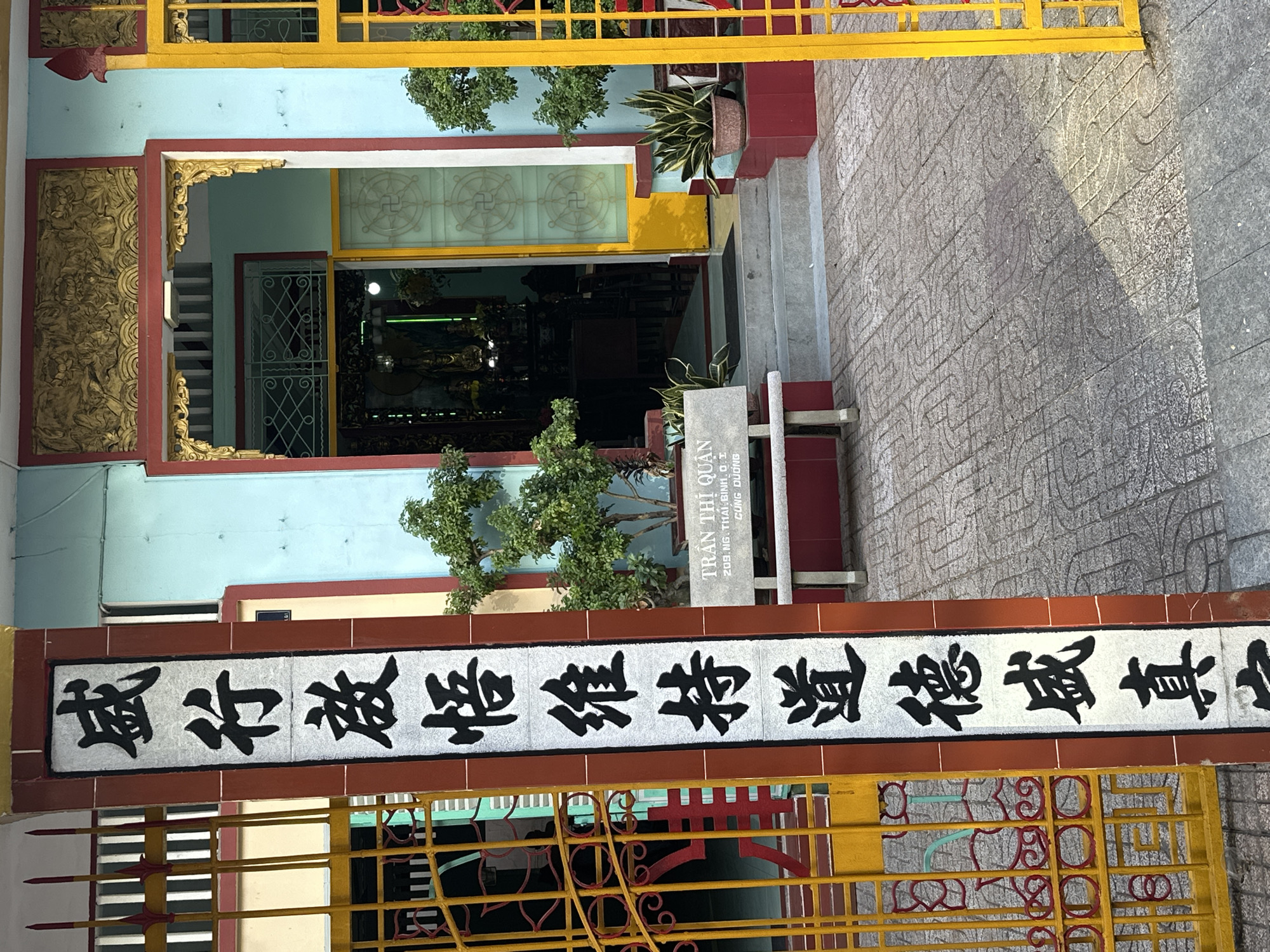 The image shows a brightly colored building, likely a temple or a place of worship, in Thủ Đức, Vietnam. The building features a light blue facade with red and yellow accents. A yellow metal gate with intricate red decorative elements is in the foreground. To the left, a gilded relief panel adorns the wall, depicting intricate patterns.
In the center of the image, an entrance to the building is visible, framed by red and gold trim. Through the entrance, the interior appears dimly lit, with glimpses of statues and possibly religious artifacts. A stone plaque near the entrance displays Vietnamese text, reading "TRẦN THỊ QUẬN 209.NG. THAI. ΒΙΝΗ.Ο.Σ CÙNG DƯƠNG". Above the entrance, a horizontal banner displays large black Chinese characters on a white background, bordered by red. The characters are stylized and appear to be a religious or philosophical phrase.
Lush green trees are planted in pots near the entrance, and more trees are visible around the building, suggesting a well-maintained outdoor space. The ground in front of the building is paved with stone tiles featuring a repeating pattern. The lighting suggests it is daytime, possibly late morning or afternoon, casting shadows on the paving stones. The overall scene conveys a sense of cultural and religious significance.