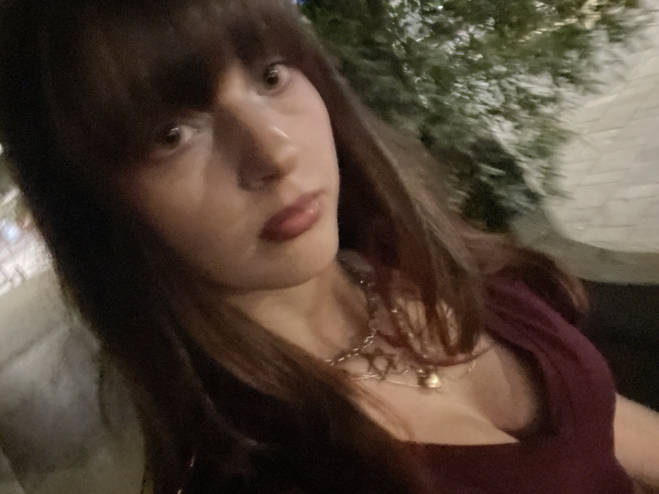 A woman with dark hair and bangs poses in an outdoor setting. She wears a dark burgundy top and a layered silver necklace. The background features blurred foliage and what appears to be a brick walkway or wall. Her expression is neutral, with dark lipstick. The lighting suggests it's either evening or a dimly lit area.