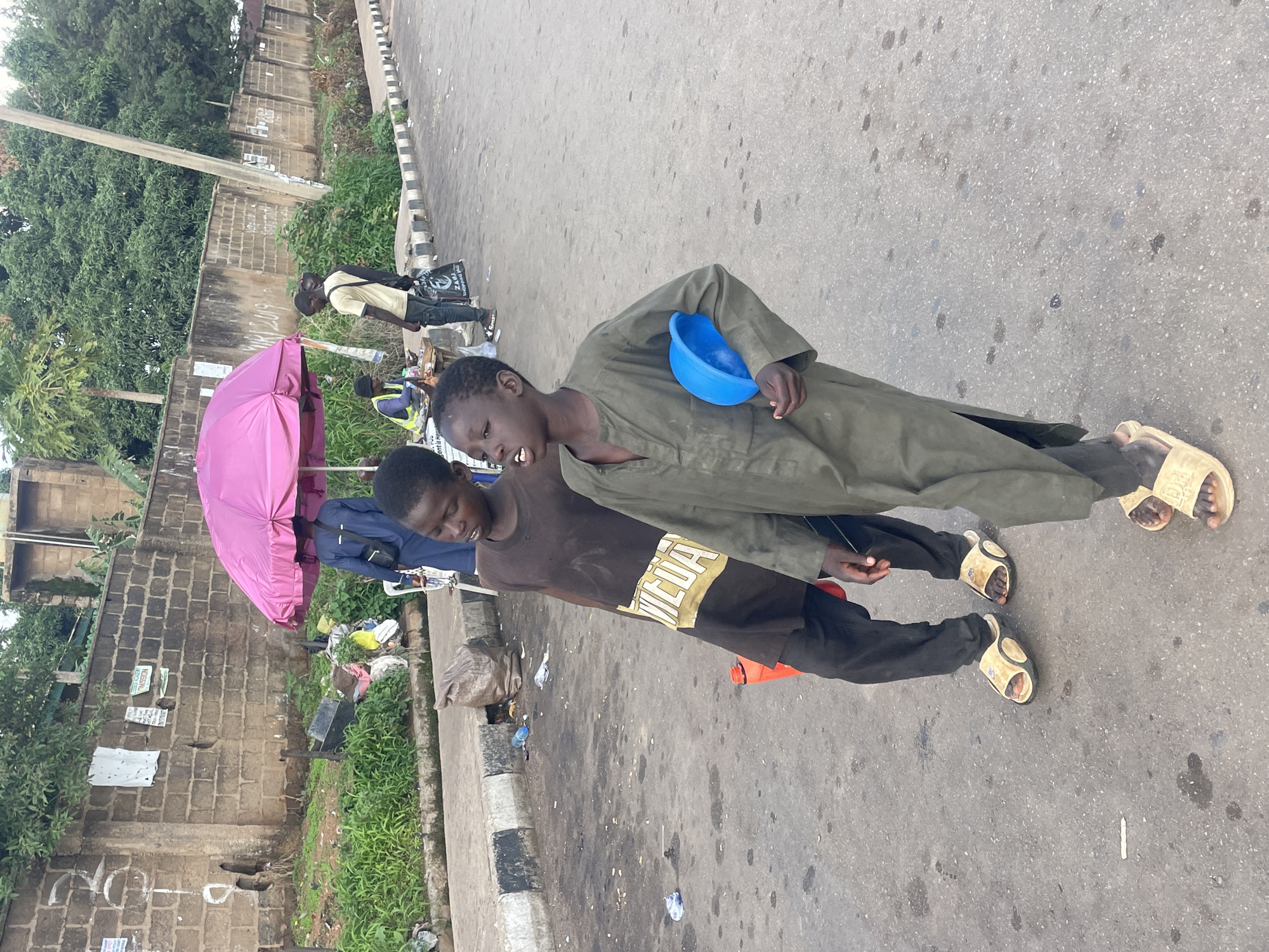 The image shows two young boys on a street in Barakin Lerawi, Nigeria. The boy in the foreground is wearing a dark brown t-shirt with a yellow and white graphic design and black pants. He is holding a blue bowl in his right hand. The boy behind him is wearing a dark green robe and black pants. Both boys are wearing light-colored sandals. They appear to be standing or walking on the side of a paved road, with a sidewalk and some greenery on the left. There is also a pink umbrella and some trash visible on the left side of the image. The overall lighting suggests it is daytime. The boys' expressions are not clearly visible, but their posture suggests they might be soliciting something or engaged in some street activity. The context of Barakin Lerawi, Nigeria, suggests an urban or semi-urban setting. There is some unreadable text on the t-shirt and a few signs on the wall behind the umbrella.