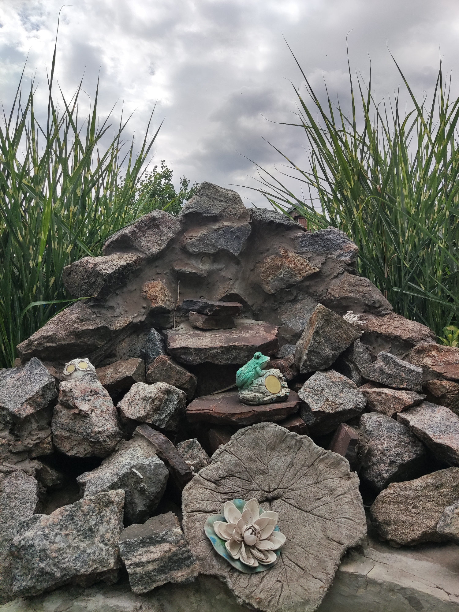 This media file depicts a garden rockery with a cloudy sky. The primary subjects are a collection of large, rough-hewn stones forming a layered structure, reminiscent of a small mountain or waterfall. In the center, perched on a reddish-brown stone, is a small, bright green ceramic frog figurine with a golden circular emblem on its base. To the left of the frog, also on a stone, is a similar golden emblem. At the bottom of the rockery, a large, flat, grey concrete slab is visible, textured to resemble a leaf. Placed on this concrete leaf is a light-colored, multi-petaled ceramic flower, possibly a lotus. Tall, green and striped grass-like plants frame the rockery on both sides, reaching towards the overcast sky. The lighting suggests it is daytime, but the heavy cloud cover indicates overcast weather. The overall impression is a decorative garden feature, possibly part of a small pond or water feature. There is no visible text in the image.