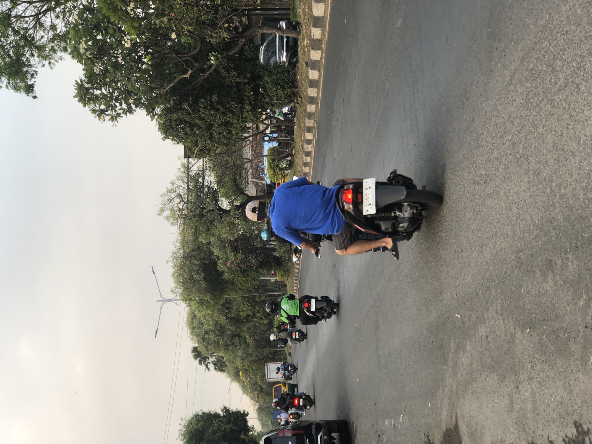 The image depicts a street scene in Ngaban, Indonesia, filled with motorcycles navigating a two-lane road. The perspective is from behind a rider on a motorcycle, capturing the road ahead. Several other motorcycles are visible in front, some with riders wearing helmets and casual clothing. One rider in the foreground wears a blue t-shirt and shorts, with a cap. To the left, a black SUV is parked or moving slowly. Trees line the right side of the road, with some buildings visible in the background. The sky is overcast, indicating a potentially hazy or cloudy day. The road surface appears to be asphalt, with painted lines marking the lanes. The overall atmosphere suggests a typical urban commute in Indonesia.