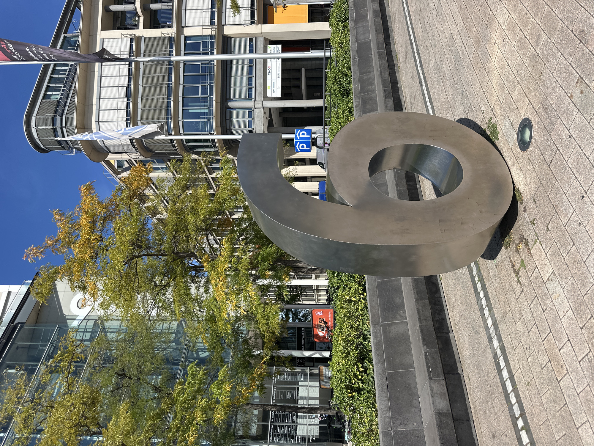 An outdoor, eye-level, full-shot photograph captures a large, brushed-metal sculpture of the number "6" positioned on a tan-and-gray paved plaza. The sculpture is in the foreground, slightly to the right of the center of the frame, with its base resting on the plaza. Behind the sculpture, a dark gray stone planter box with a matching stone bench runs horizontally across the midground. Beyond the planter, a row of manicured green bushes lines the base of modern buildings.
The buildings are multi-story structures with glass facades, visible balconies, and slatted window coverings. To the left, a building with a large glass section reflects the sky and trees. To the right, a building with a curved facade features multiple floors with numerous windows. Two flags are visible on poles in front of the building on the right: one is white with blue stripes, and the other is dark with text. A blue and white sign indicating parking ("PP") is mounted on a pole behind the sculpture, on the left side of the frame. Trees with yellowing leaves, suggesting autumn, are interspersed between the sculpture and the buildings, casting dappled shadows on the plaza. The sky is a clear, bright blue. A small, circular ground light is visible in the lower right quadrant of the image, set into the paving stones.
The scene is located in Cologne, Germany, specifically in the MediaPark complex. The sculpture is positioned in front of the "Cologne Medienpark" building.