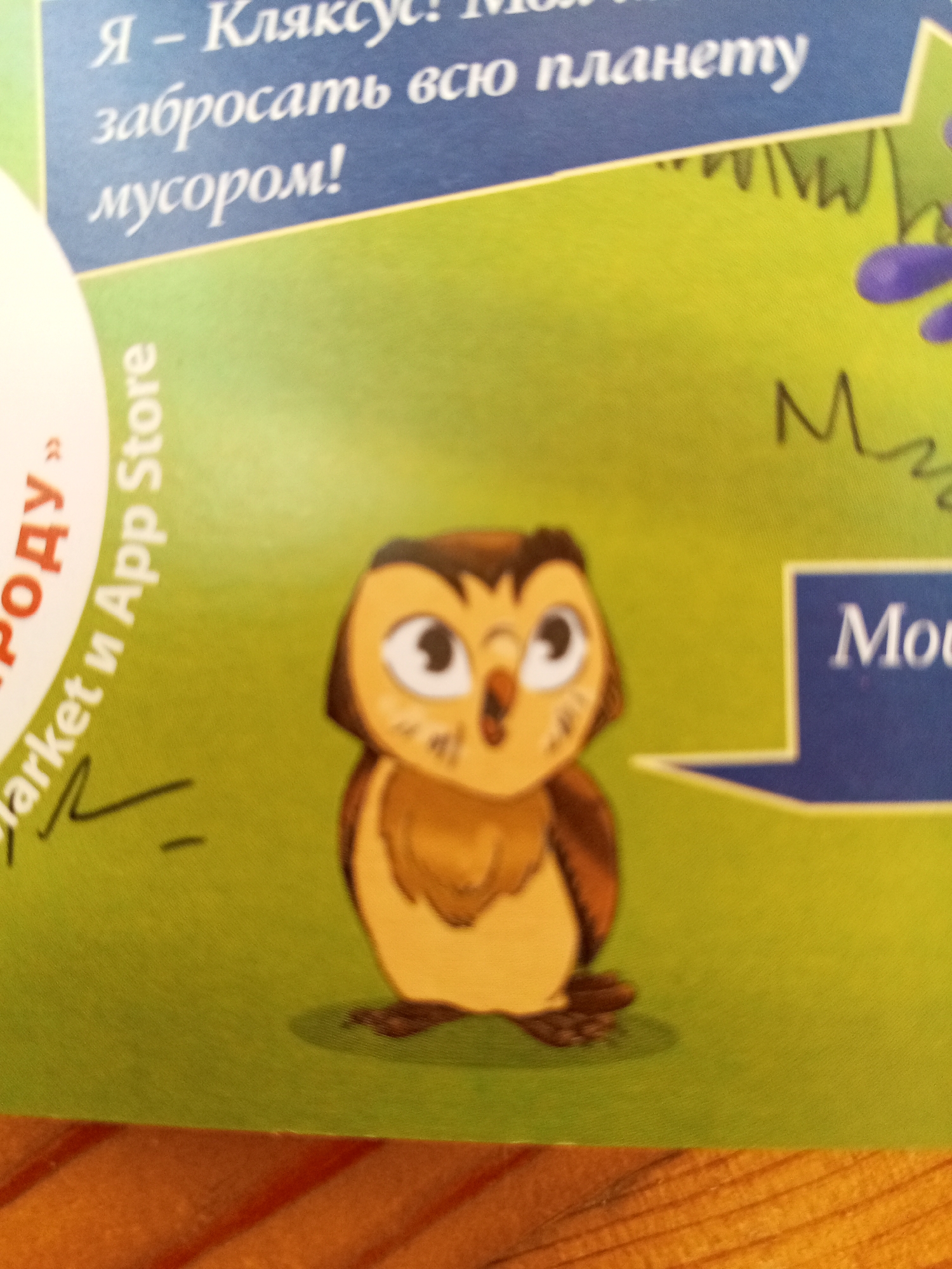 The image shows a close-up of a cartoon owl on a green surface, with Russian text in blue and white in the upper left corner, and text within a blue speech bubble in the lower right. The Russian text in the upper left reads "Я – Кляксус! Моя планета", followed by "забросать всю планету мусором!". The text within the blue speech bubble reads "Mou". There are also some stylized drawing elements on the green surface, including what appears to be a branch or vine and a series of 'M' shapes. The lower portion of the image shows the edge of a wooden surface. The image is a photograph of printed material. There are no people or animals present, other than the cartoon owl. The location in Chistopol, Russia, is not ascertainable from the provided image.