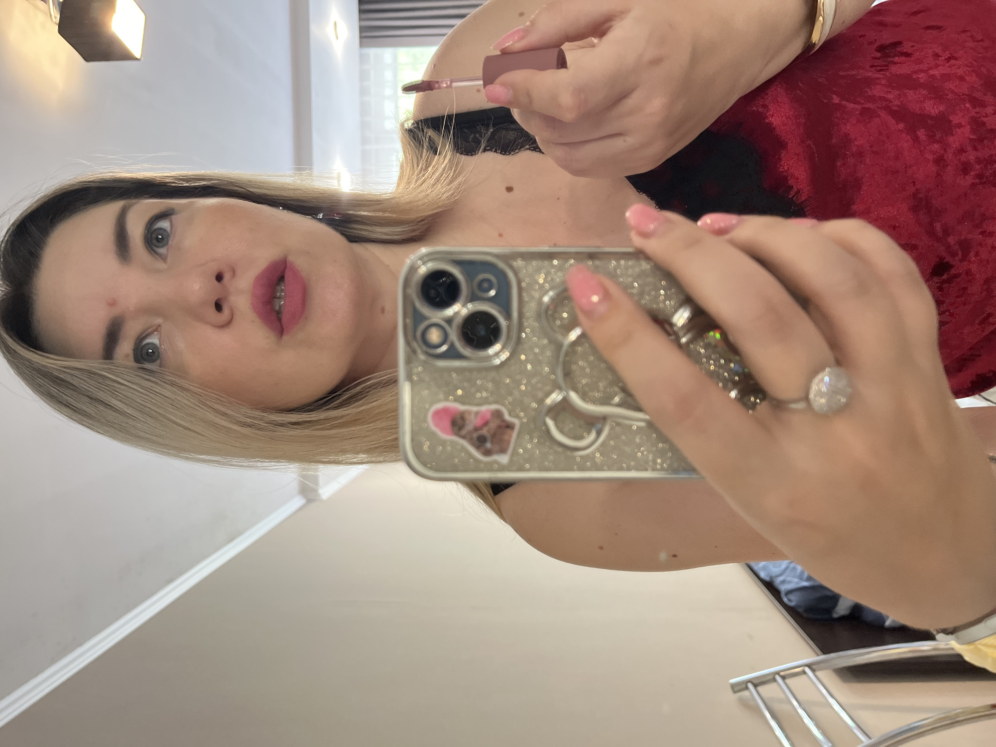 A woman with blonde hair and pink lipstick is taking a selfie in a room. She is wearing a red velvet camisole with black lace trim. She is holding a phone in her left hand, which has a sparkly case with a cartoon sticker on it. In her right hand, she is holding a tube of lip gloss. She is wearing a large diamond ring on her left ring finger and a bracelet on her right wrist. The room appears to be a bedroom or dressing area, with a mirror, a light fixture on the ceiling, and a metal chair visible. There are no specific indicators that place the location in Sochi, Russia.