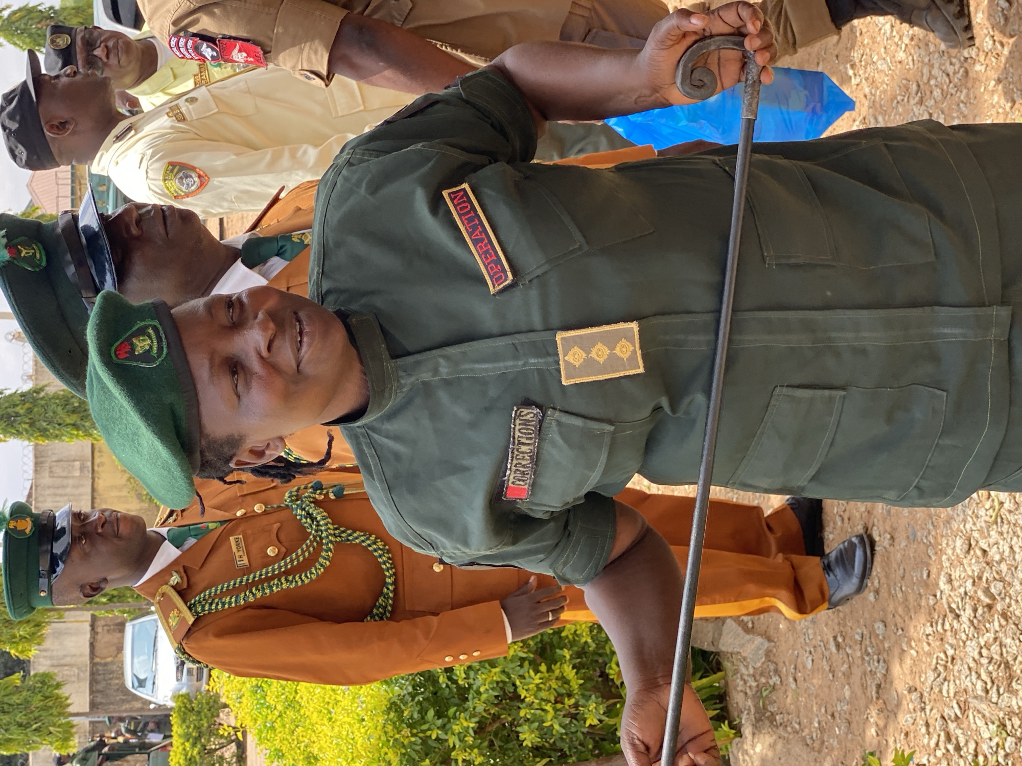 The image shows a group of people, likely correctional officers, in uniform. The primary subject is a man in the foreground wearing a dark green uniform with a green beret. He is holding a black baton-like object with a decorative metal handle. Patches on his uniform read "OPERATION" and "CORRECTIONS". He appears to be looking slightly up and to the left, with a neutral expression.
Behind him, other individuals are visible in various uniforms. To his left, a man in a lighter brown uniform with a green and yellow cord across his chest looks towards the camera with a slight smile. Another man, further back and to the left, wears a light yellow uniform and looks upwards. To the right of the primary subject, a person in a brown uniform is partially visible, also holding a similar baton. The setting appears to be outdoors, with gravel on the ground and some greenery in the background. The lighting suggests it is daytime, possibly morning or afternoon, with mild weather. The presence of uniforms and official-looking attire points to a formal event or duty. The image is oriented vertically.