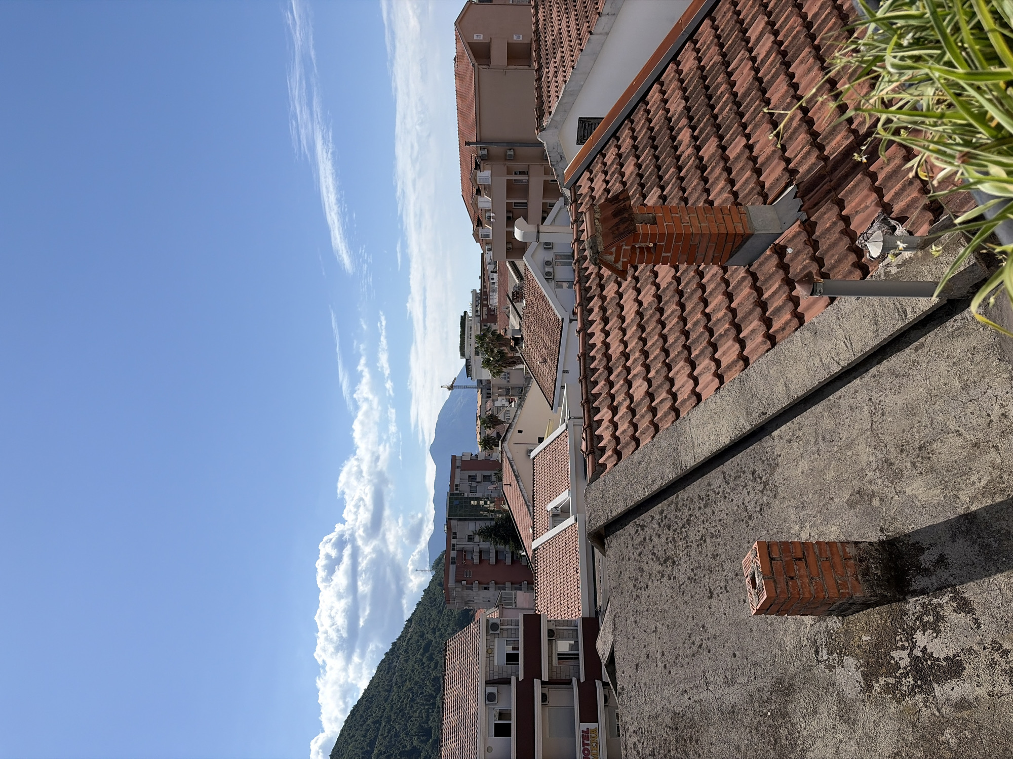 The image, when viewed in its correct orientation, provides a sunlit, elevated perspective over a cityscape in Budva, Montenegro, dominated by traditional red-tiled roofs.
In the immediate foreground, a textured concrete wall runs horizontally, featuring a small, hollow brick structure protruding from its surface, casting a distinct shadow. Above this wall, a section of a red-tiled roof is visible, with a green and white variegated plant cascading over its edge. A brick chimney also rises from this roof.
The midground is filled with numerous multi-story buildings, characterized by their terracotta roof tiles and light-toned facades. Many buildings display balconies and external air conditioning units. A partial sign on one building clearly shows the text "HOTEL MENA".
The background reveals a natural landscape, with a large, verdant, tree-covered mountain ascending on the left side of the frame. Further in the distance, a bare, grey mountain peak can be seen under the clear sky. The sky itself is a vibrant blue, adorned with sparse, wispy white clouds stretching across the upper half and more substantial cumulus clouds gathered closer to the mountains. A faint outline of a construction crane is visible near the distant mountain.
The scene is bathed in bright daylight, indicated by the crisp shadows cast by structures and foliage. No human activity or specific events are depicted, presenting a static, picturesque view of the urban and natural environment.