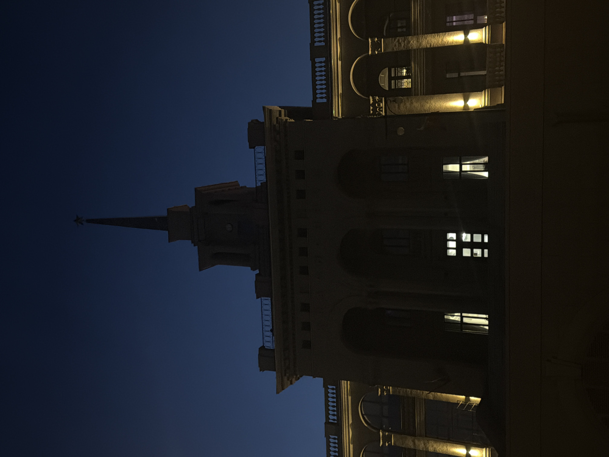 The image displays a monumental building against a deep blue, post-sunset or night sky. The architecture is grand and imposing, strongly resembling the Stalinist Empire style prevalent in Russia, and is identifiable as the Krasnoyarsk Regional Museum in Krasnoyarsk, Russia.
A tall, multi-tiered spire, topped with a distinctive star-like ornament, dominates the upper part of the structure. The building's facade features elaborate architectural details, including large arched windows, decorative pilasters, cornices, and balconies with ornate balustrades. Warm yellow lights illuminate various sections of the building, highlighting its intricate design and contrasting with the darker, unlit portions and the dark blue sky. Lights are also visible inside some windows, indicating interior activity or general illumination.
The scene conveys a sense of quiet majesty, with no people visible and no specific event taking place beyond the building being lit up for the night. The dark sky confirms the time of day as evening or night. The image's original orientation, rotated ninety degrees, emphasizes the building's verticality and soaring presence against the sky. No visible text is present within the frame.