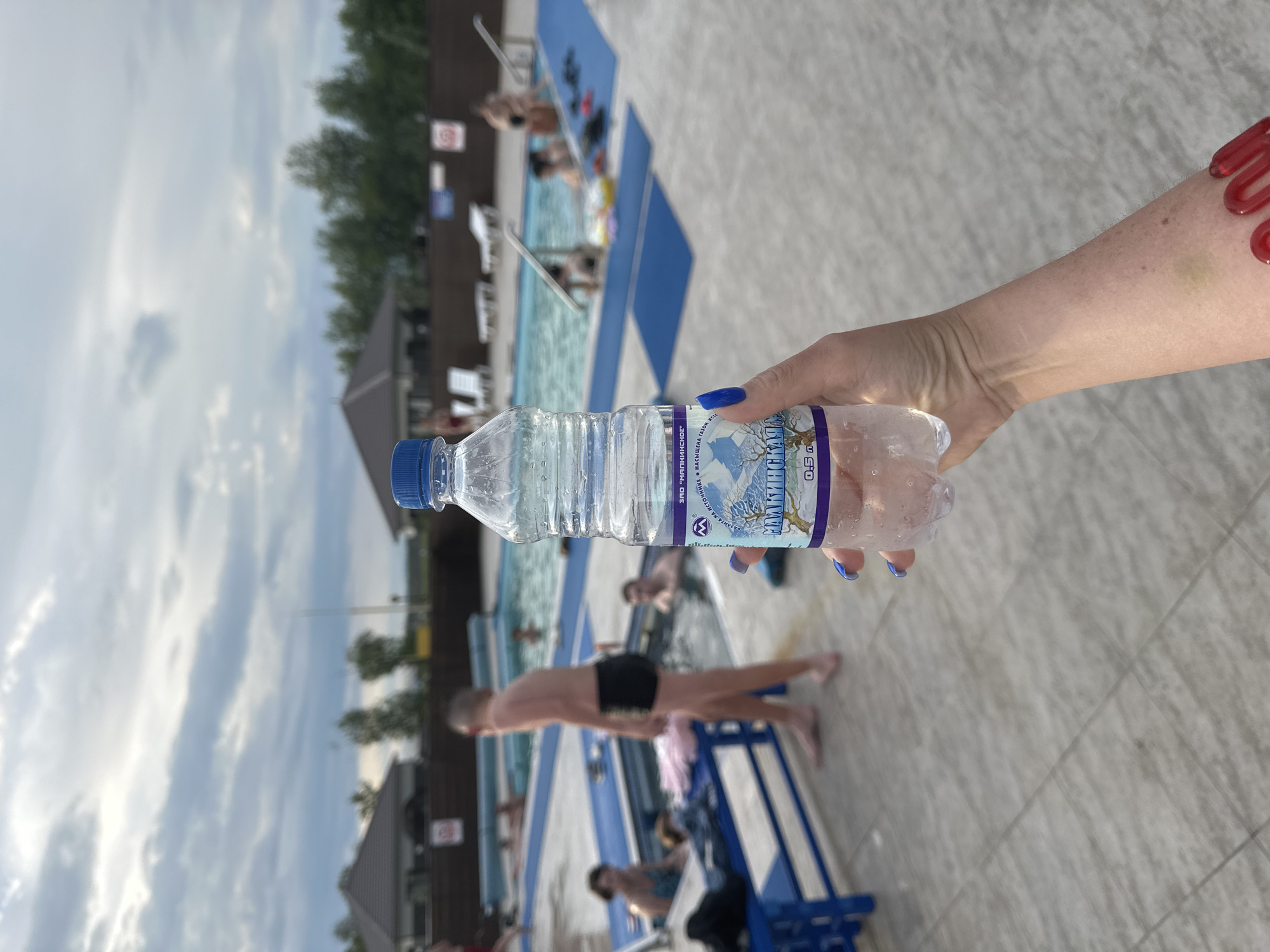 In Yuzhnyye Koryaki, Russia, a person holds a bottle of mineral water in the foreground, with a swimming pool and surrounding amenities visible in the background. The pool area features blue mats and lounge chairs, and several people are in and around the water. The sky above is overcast. The image captures a leisure scene at an outdoor pool.